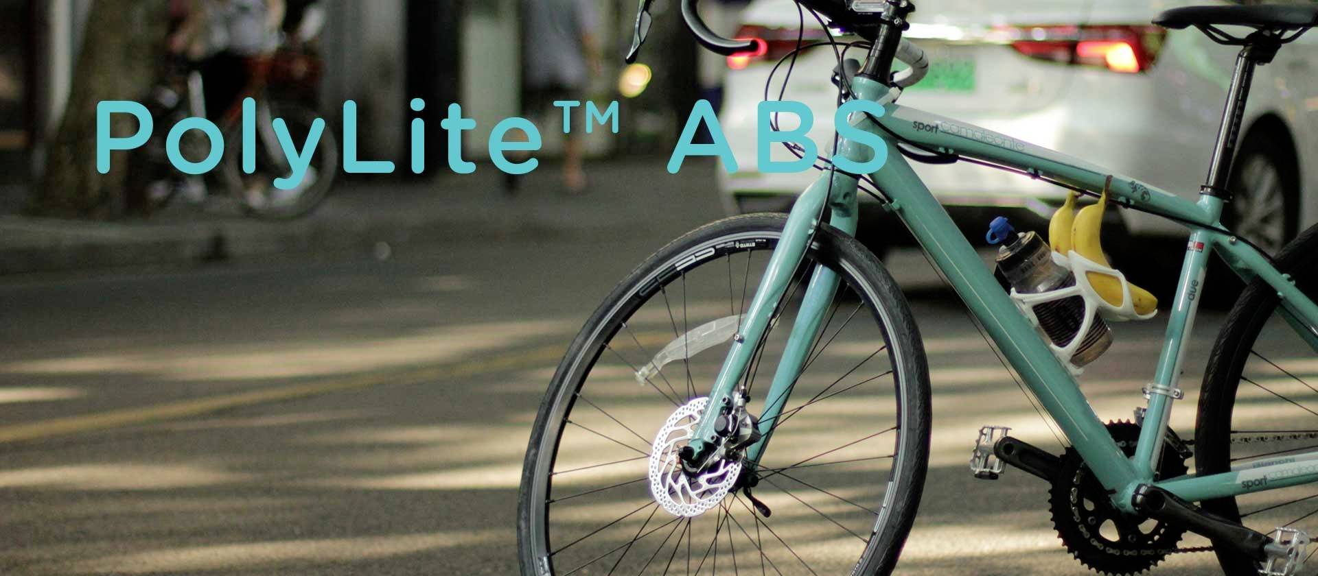 A turquoise bicycle is parked at the roadside with a 'PolyLite ABS' inscription in the background of the image. Cars can be seen in the background.