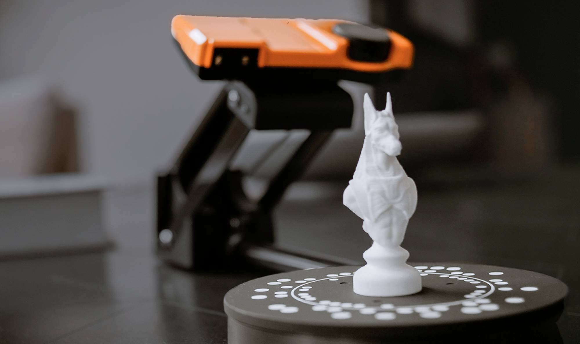 The image shows a white chess knight on a round table with a 3D scanner behind it. The scanner is orange and black.
