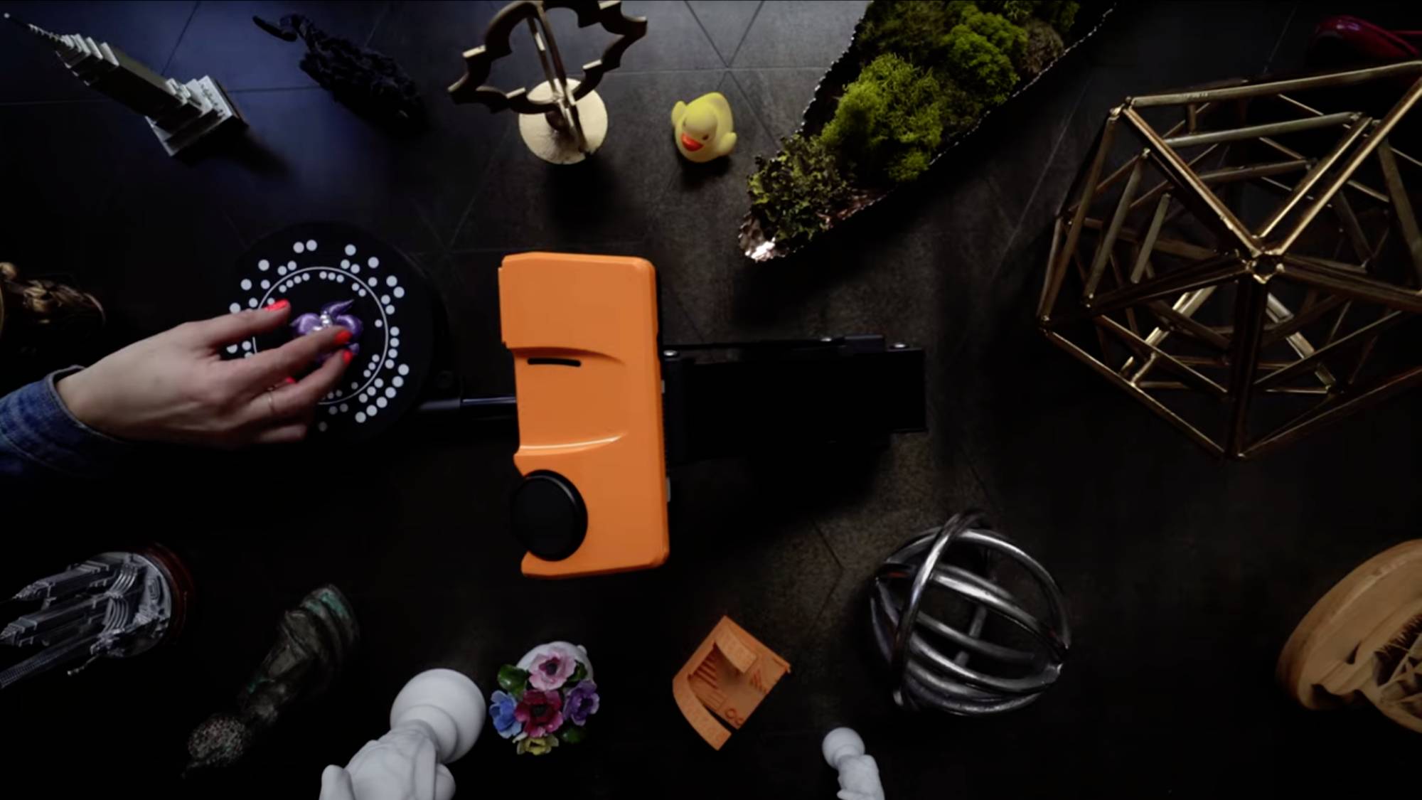 A person is turning a knob on an orange device, surrounded by decorative objects such as figurines and geometric shapes on a table.