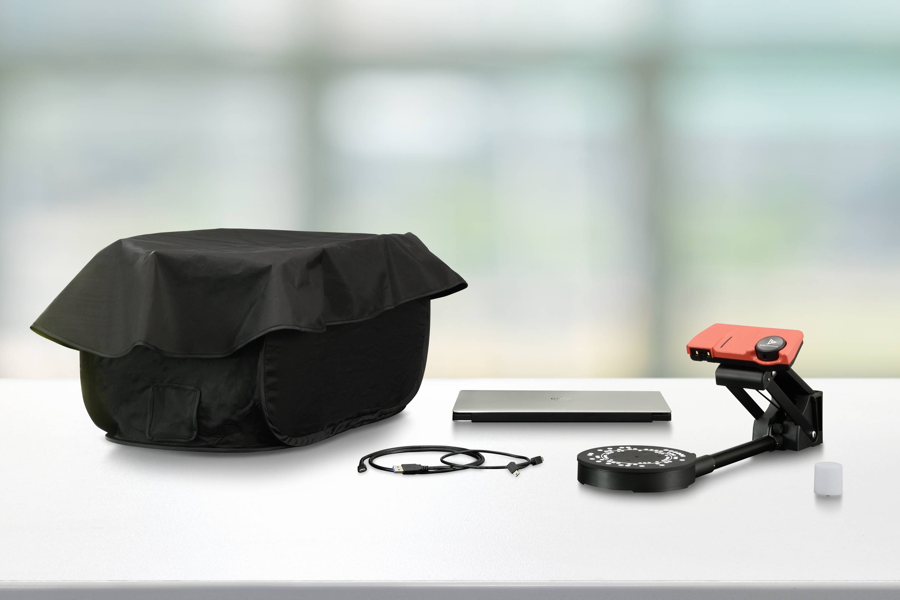 A laptop, a projector with a red cover, and a black protective cover on a table in a bright room.