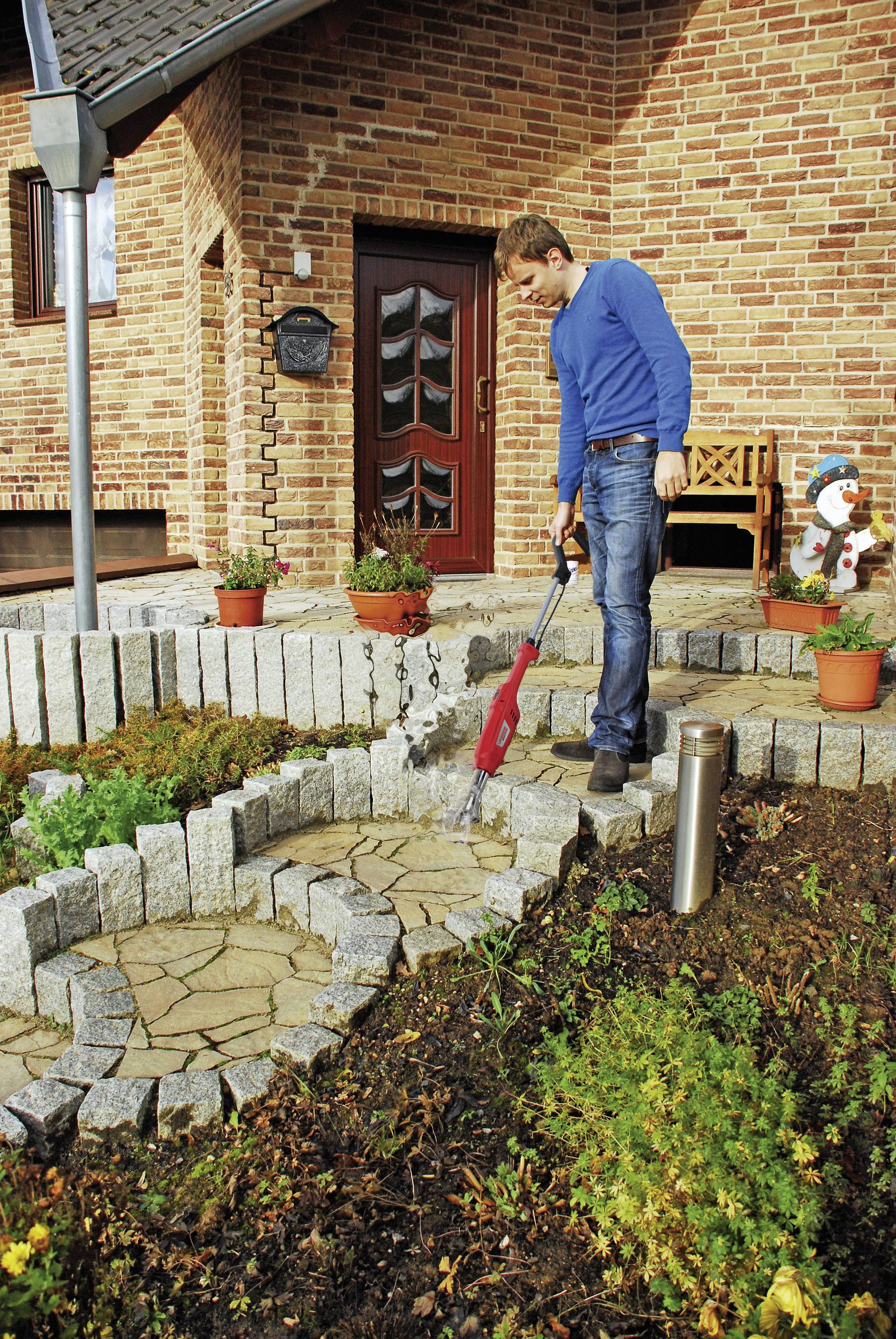A man in blue clothing is cleaning a stone staircase with a leaf blower in front of a house. The scene depicts a sunny autumn day.