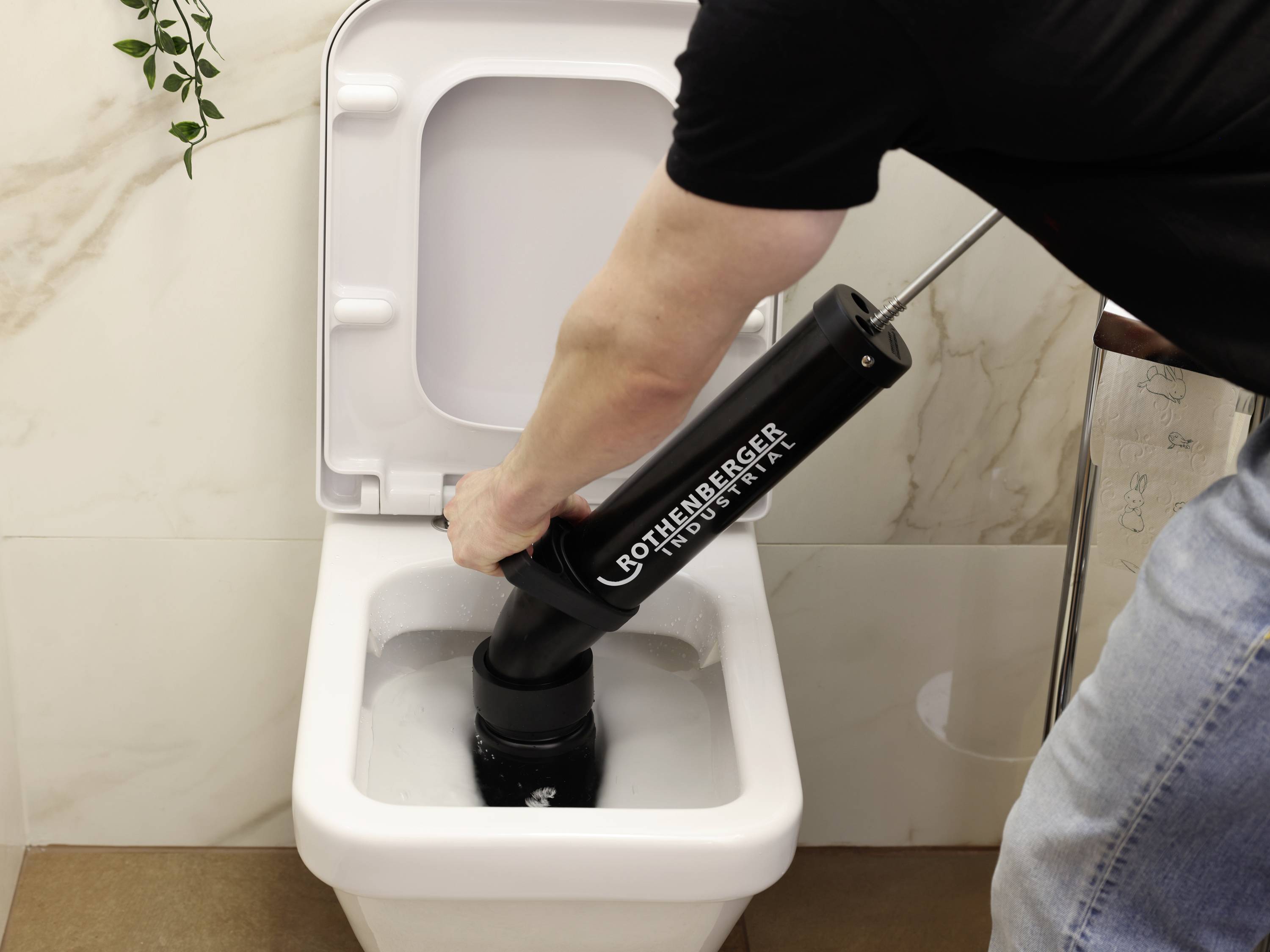 A person is using drain cleaner to unblock a toilet in a bathroom. A plant is hanging on the wall.