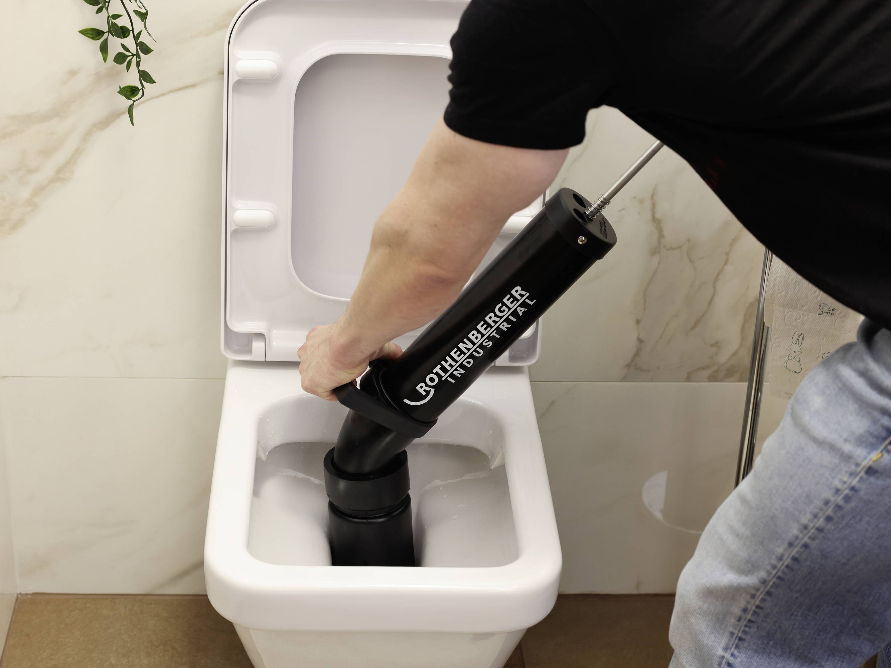 A person is using a large tool to unblock a toilet. The tool is black and is held over the toilet bowl.