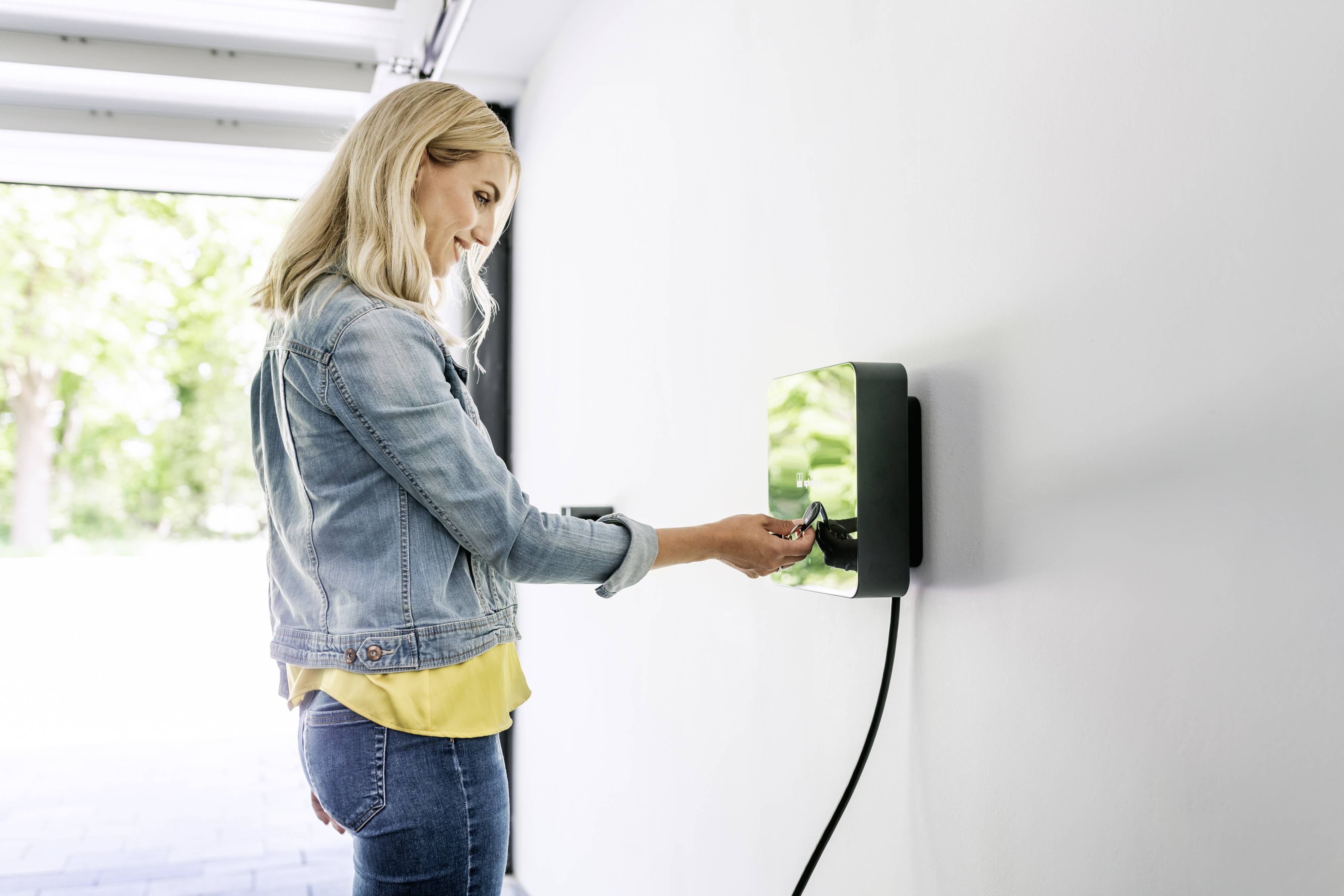 A woman is charging an electric vehicle at a wall-mounted charging point. She is wearing a denim jacket and standing in an open garage.