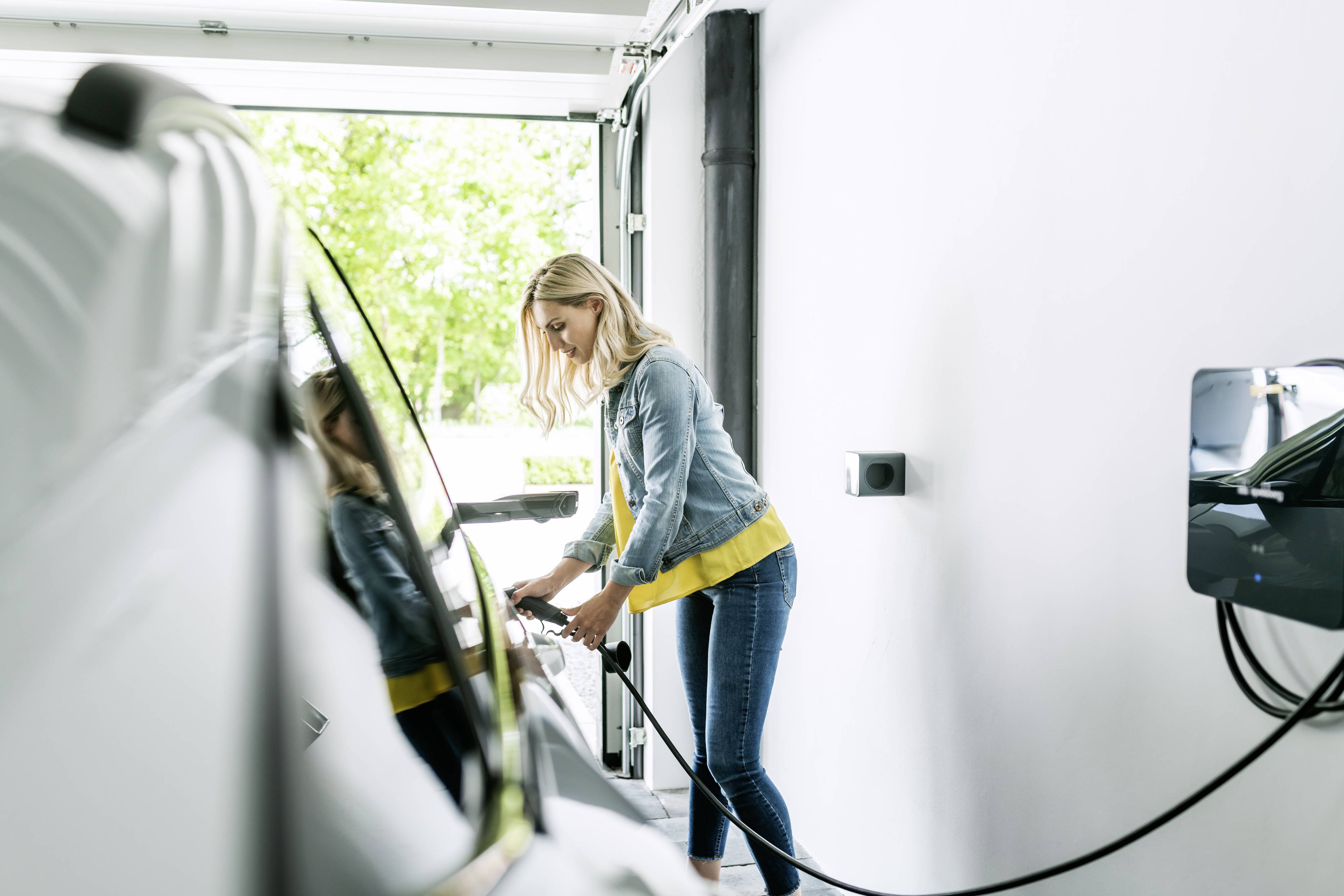 A woman is charging an electric car in a garage. She is holding the charging cable while standing next to the vehicle. The garage is brightly lit.