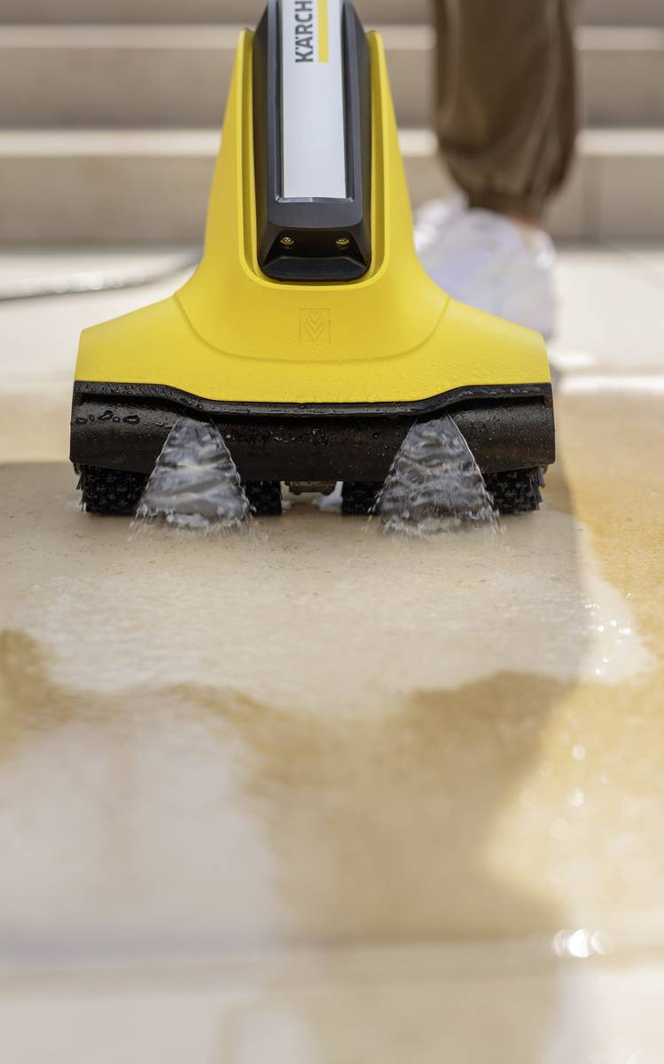 A yellow and black pressure washer sprays water onto a stone floor, with a person wearing trainers standing nearby.
