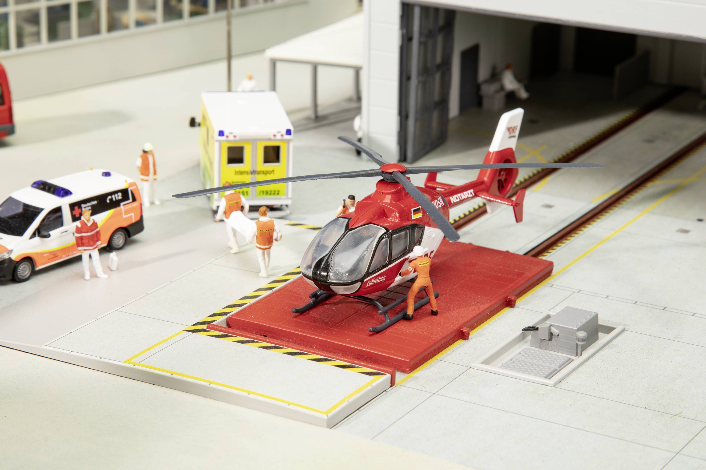 A model of a helicopter on a landing platform with figures wearing rescue clothing, an ambulance, and a medical vehicle in the background.