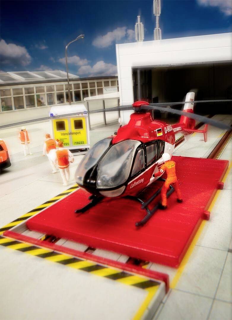 A red rescue helicopter is parked on a platform. Surrounded by several figures in orange overalls. A ambulance is in the background.