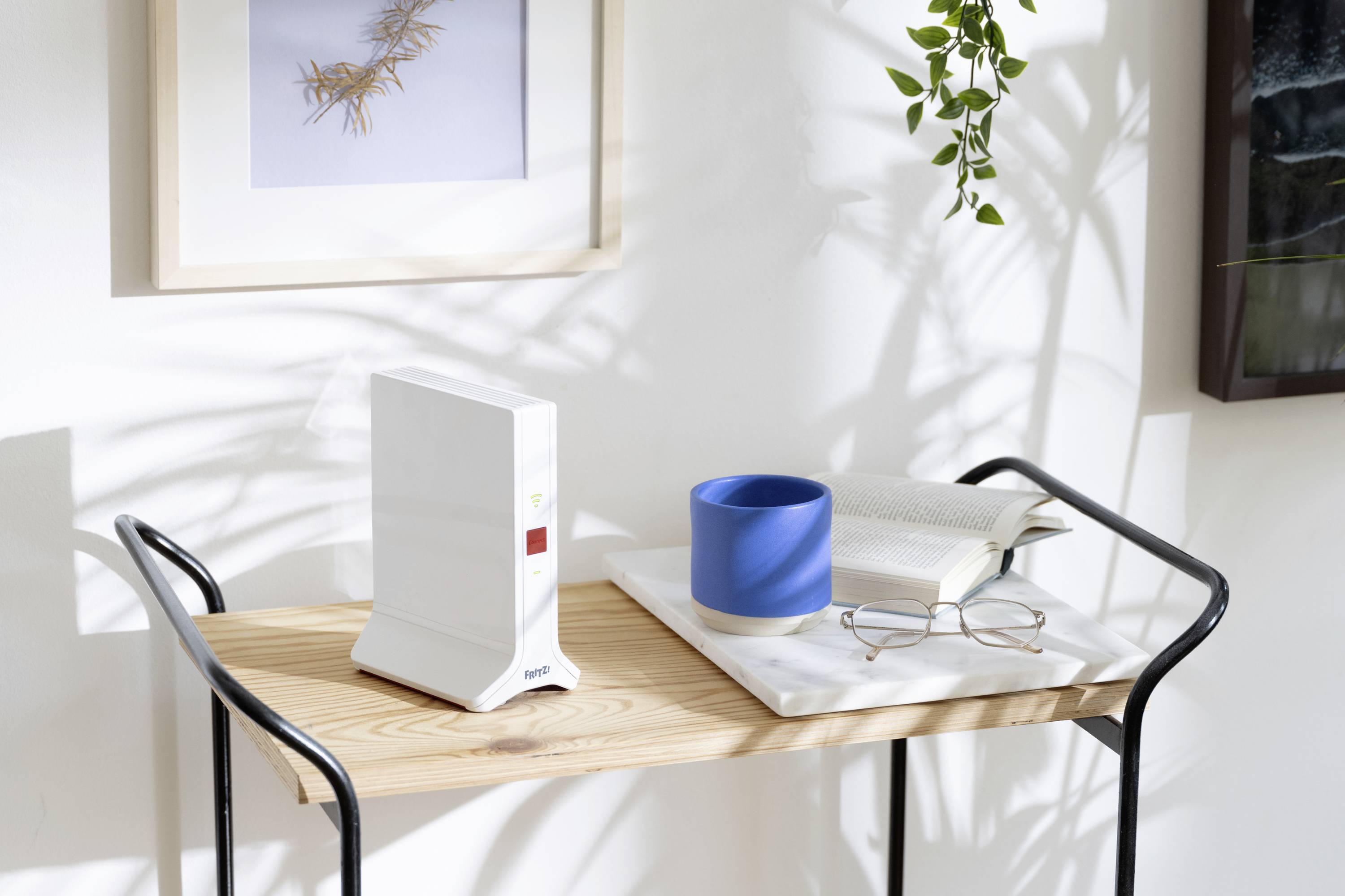A router on a wooden table beside a blue mug, book and glasses, surrounded by plants in sunlight, creates a cosy atmosphere.