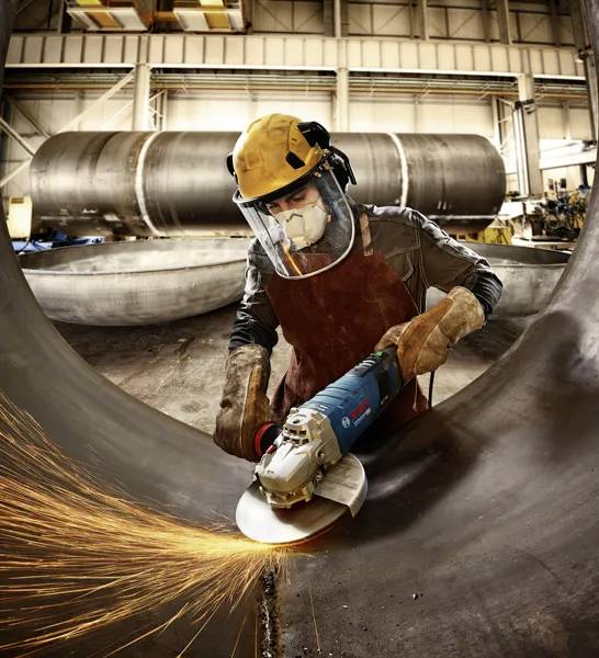 Worker in safety gear uses a grinder on large metal pipe, sparks flying, in a spacious industrial workshop with cylindrical tanks.