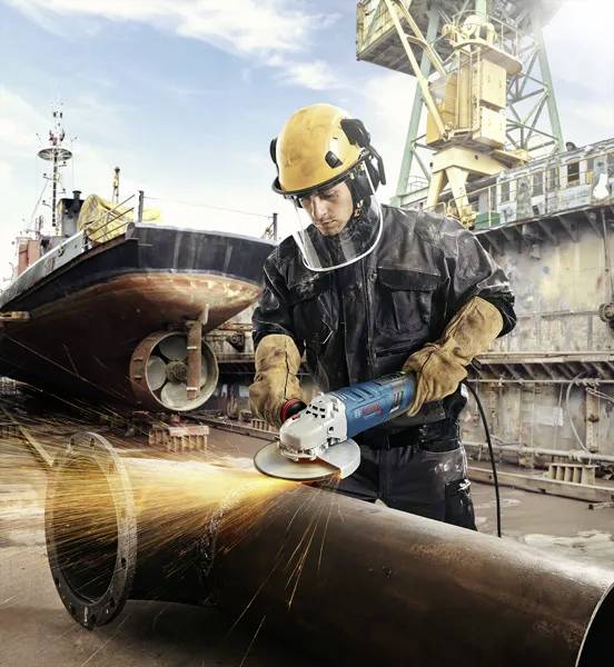 A worker in safety gear grinds a metal pipe, emitting sparks, with a ship and crane in the background at a shipyard.