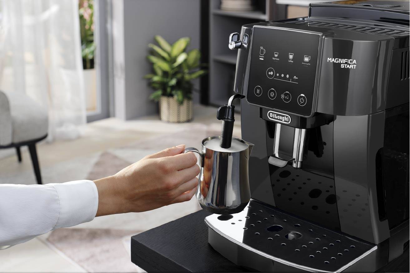 A person is frothing milk using an espresso machine. The kitchen is modern in design, with plants in the background.