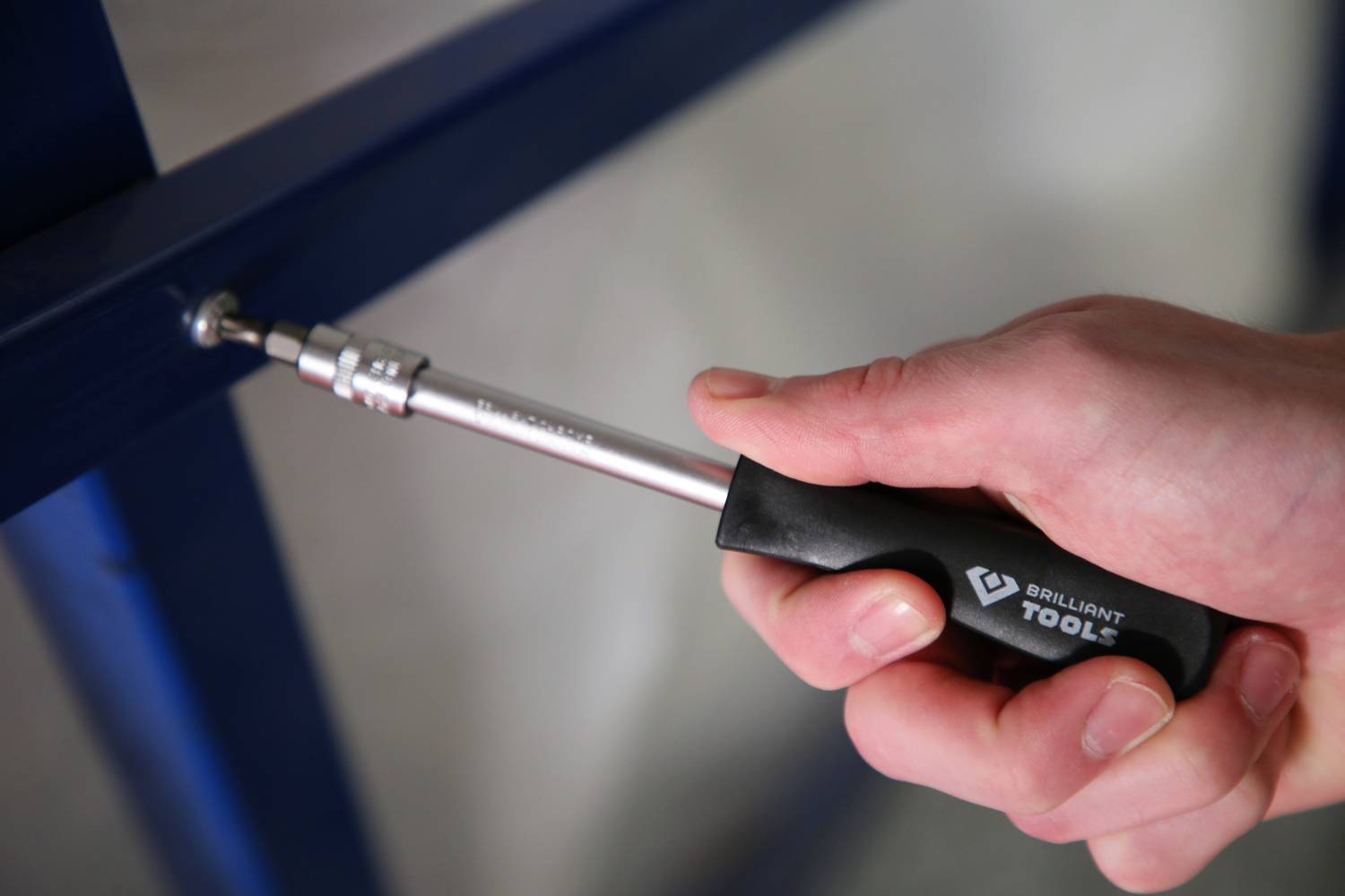 A hand is holding a screwdriver tightening a screw in a blue metal frame. Brand name visible on the handle.