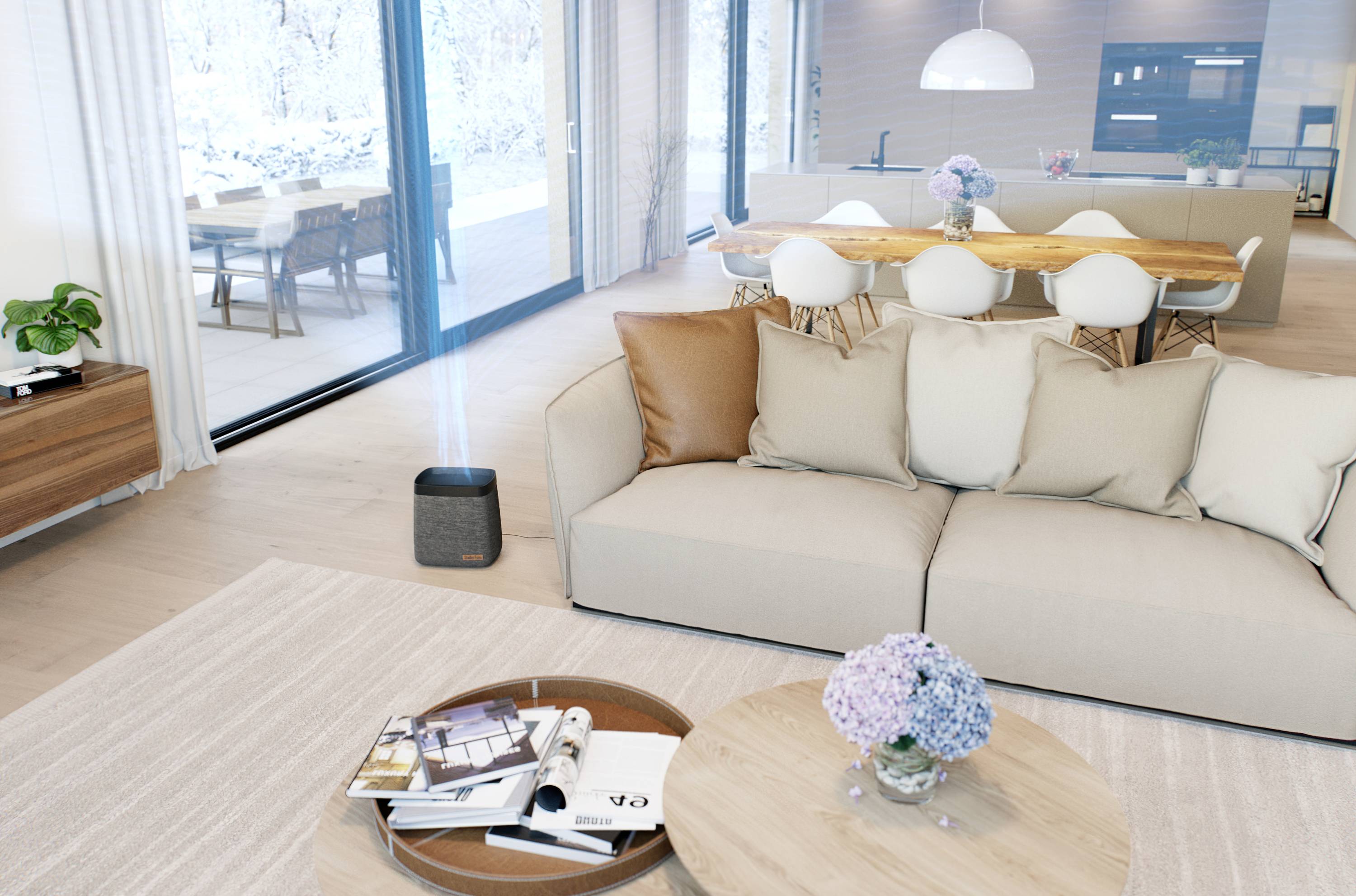 A modern living room with a sofa and a coffee table, featuring books and flowers, with a dining area and large windows in the background.