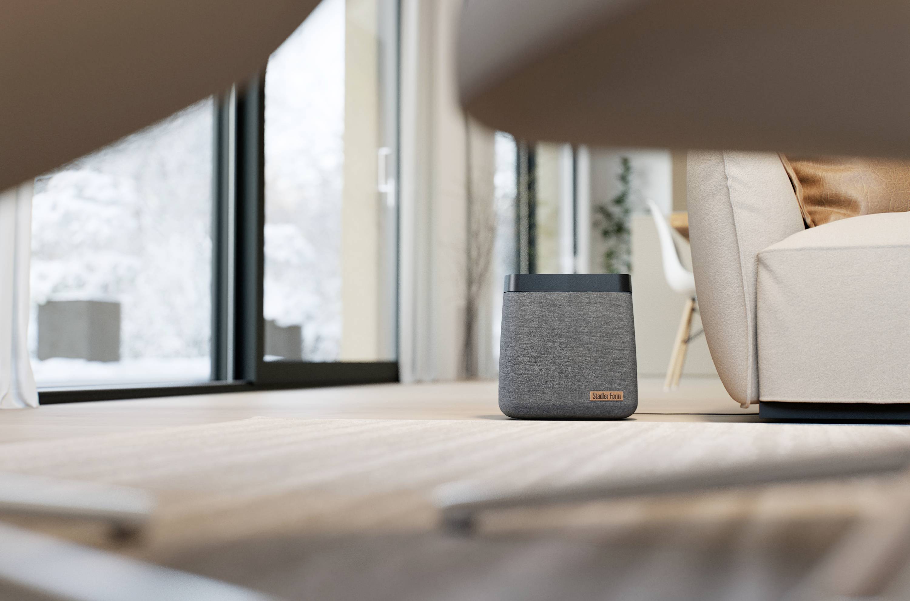 A grey speaker sits on a wooden floor in a modern living room, with large windows overlooking a snow-covered landscape.