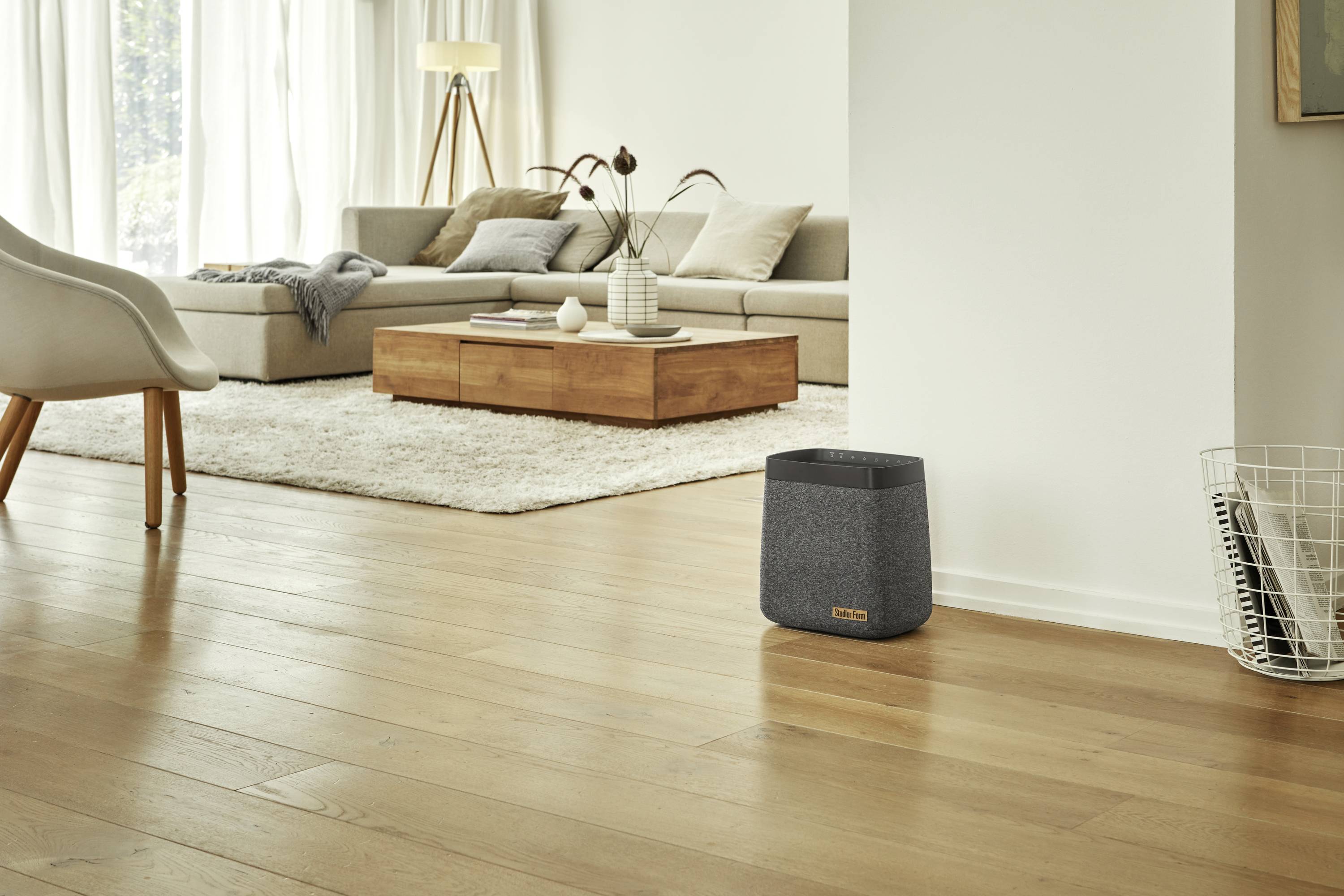 Living room with sofa, wooden coffee table, light-coloured rug, armchair, standard lamp and a grey air purifier on the wooden floor.