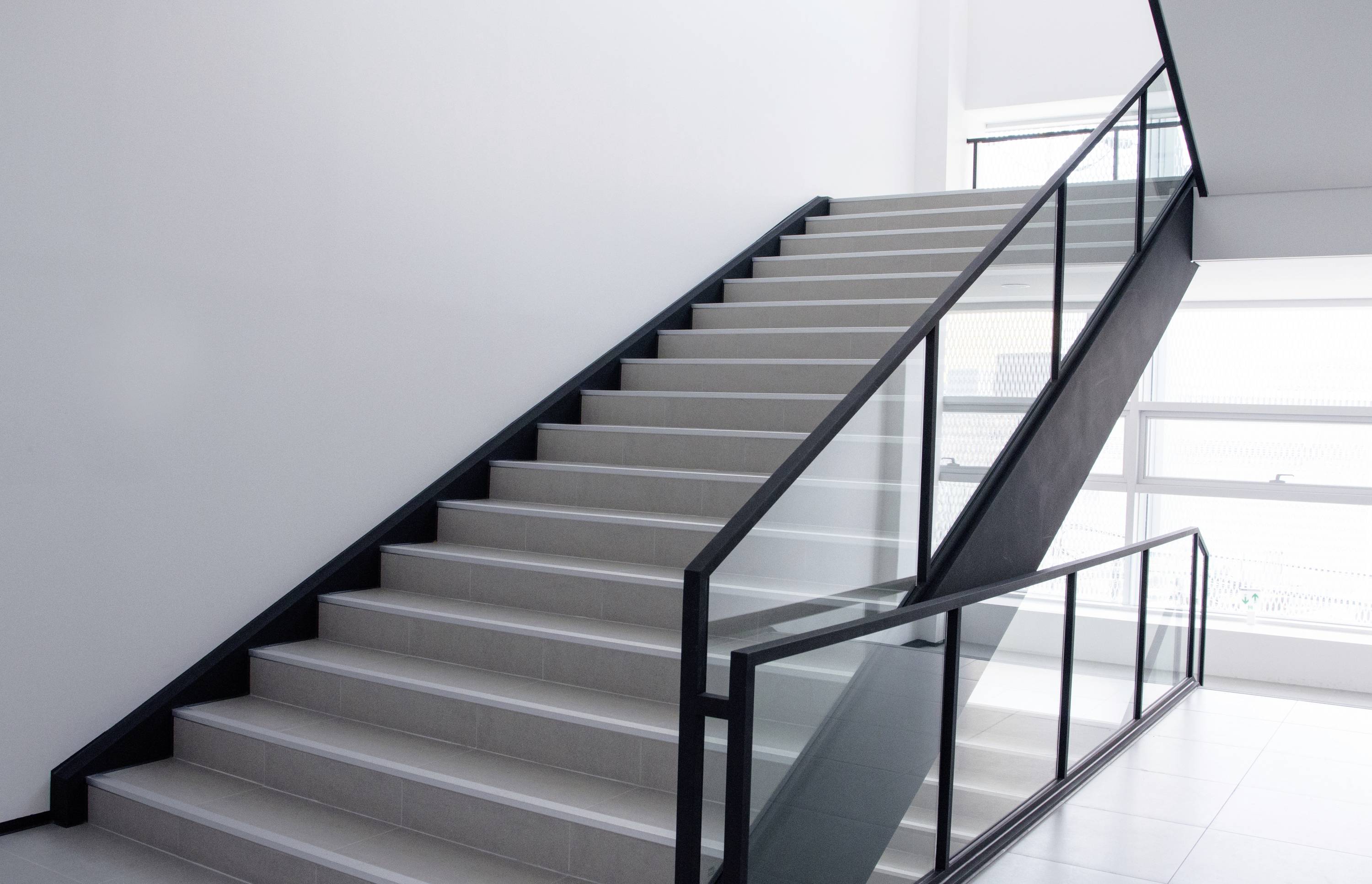 A contemporary staircase with a black handrail and glass balustrade in a bright, minimalist building. Natural daylight floods through large windows.