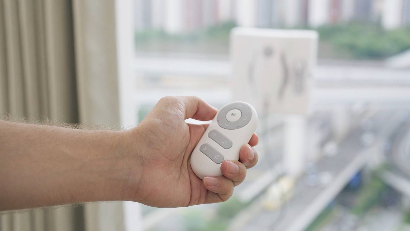 A hand is holding a white remote control near a window. Outside, a street and buildings can be seen blurrily in the background.