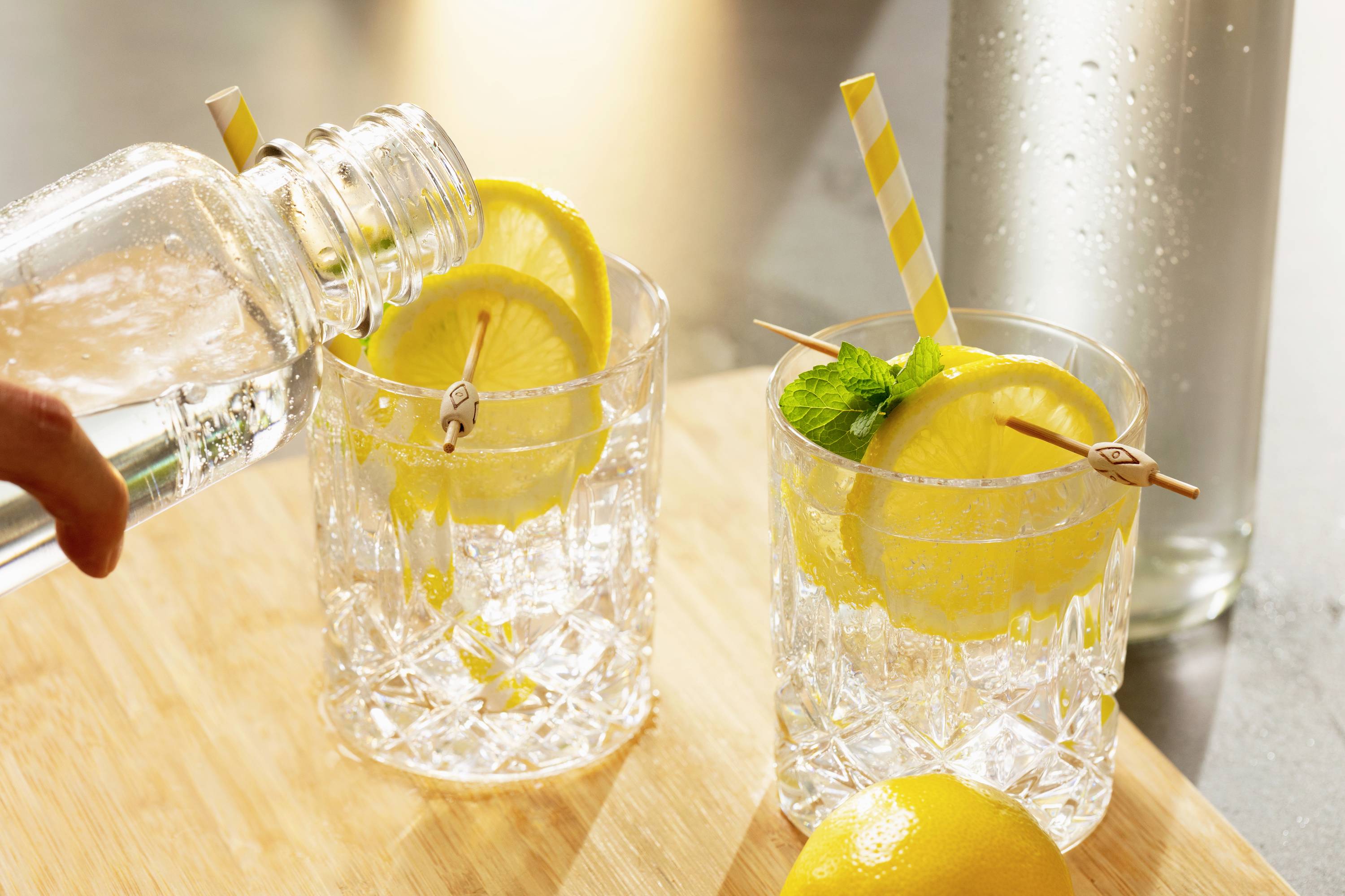 A glass of water with ice, a slice of lemon and a mint leaf is being poured. A second glass stands beside it on a wooden board.