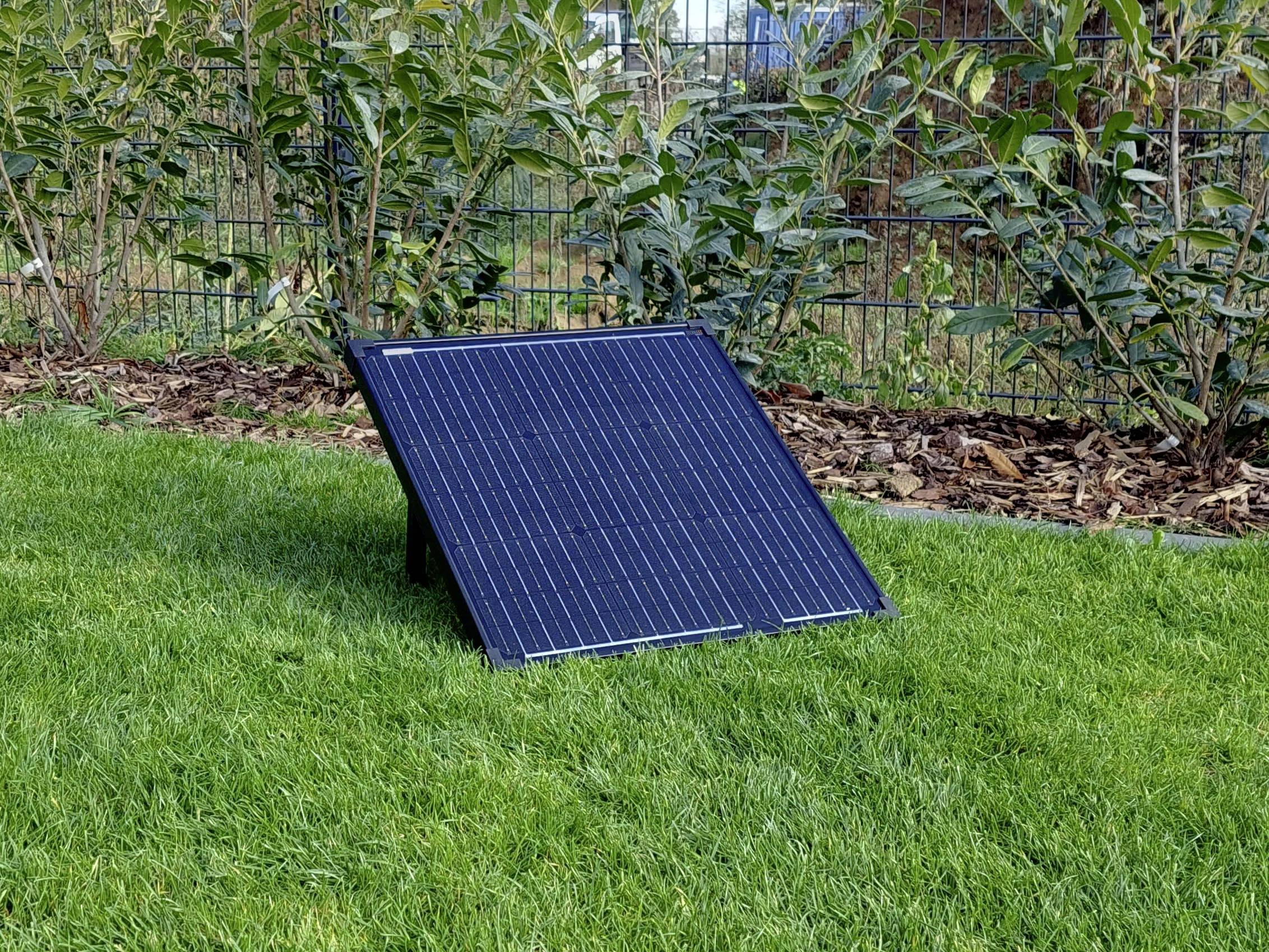A solar panel stands at an angle on the green lawn in a garden. Plants and a fence are visible in the background.
