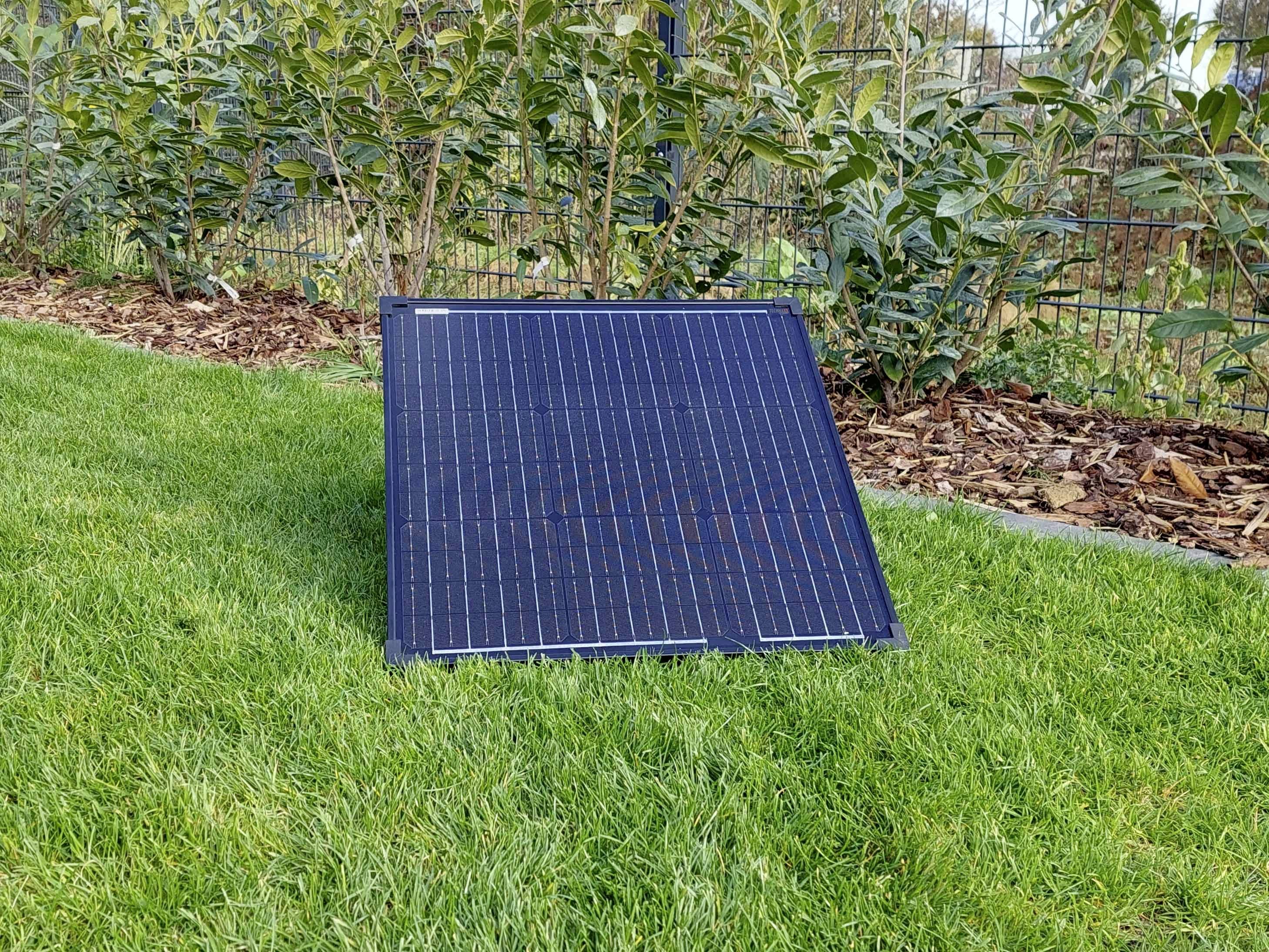A solar panel stands upright on a grassy area, surrounded by low shrubs.