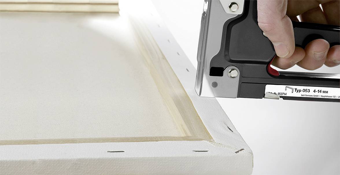 A hand uses a staple gun to secure canvas fabric to a wooden frame. The fabric is stretched taut at the corners.