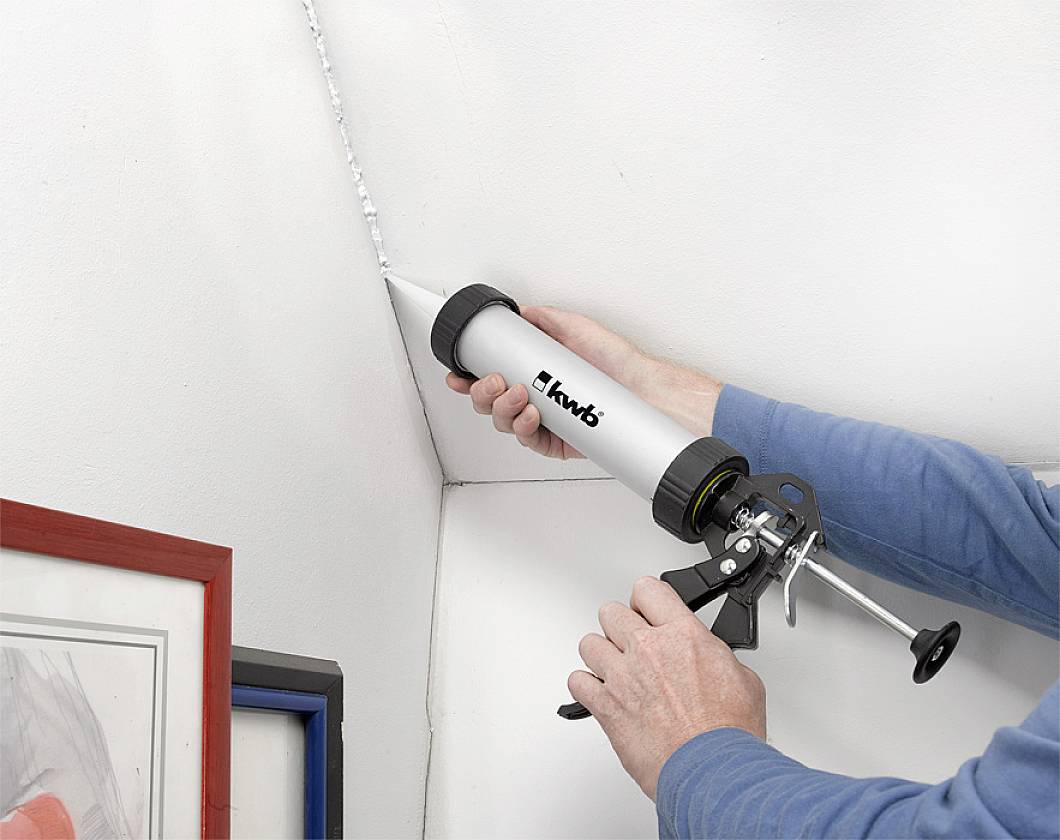A person is using a cartridge gun to apply a white sealant into a corner between two walls.