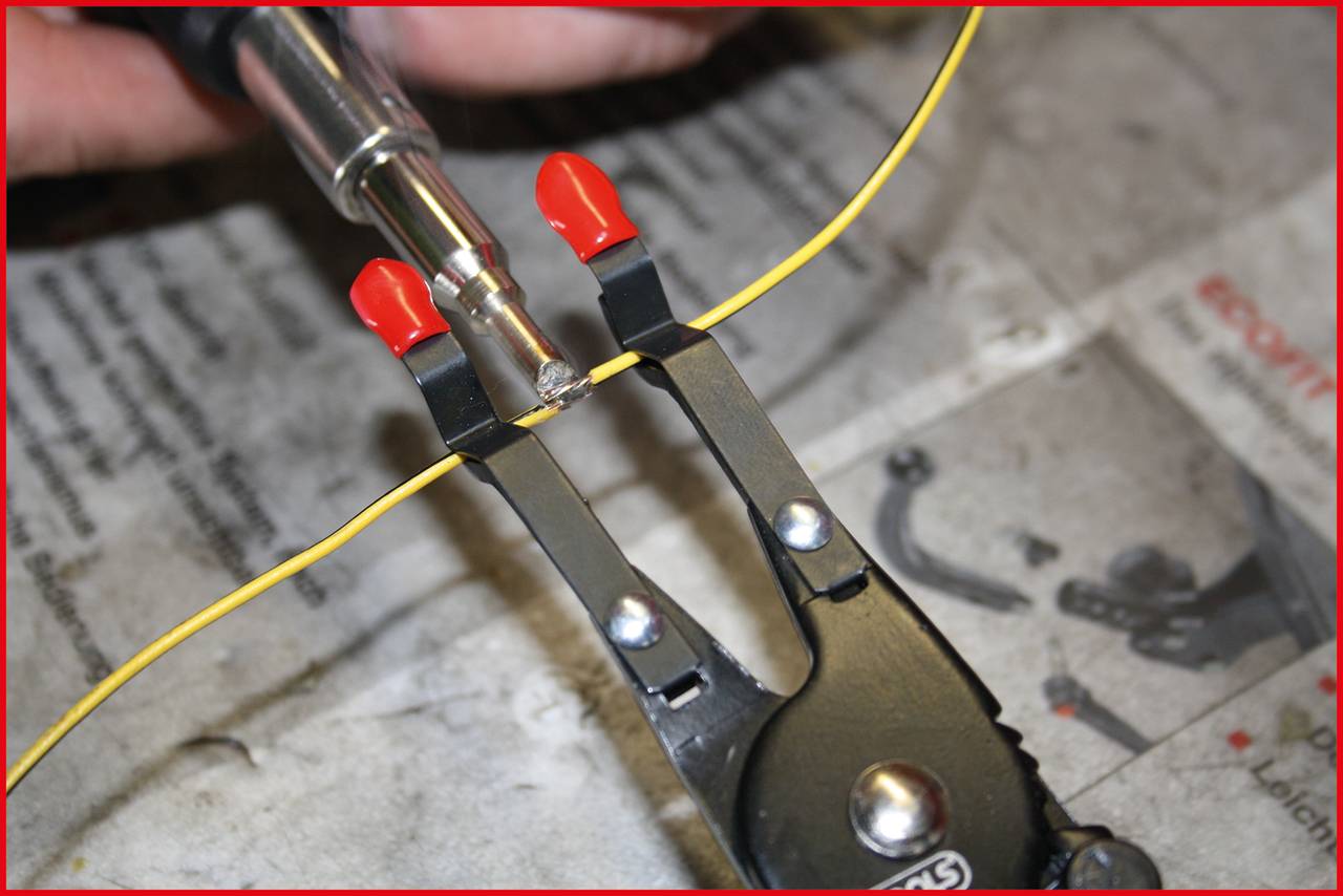 Close-up of a yellow electrical wire being held with pliers while a soldering iron heats its end. The background is a newspaper.