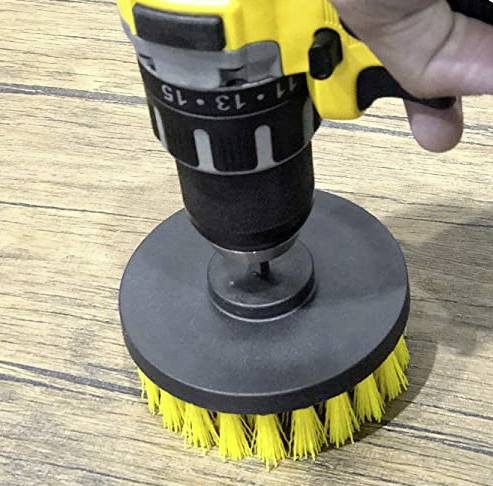An electric drill with a yellow brush attachment is cleaning a wooden surface. A hand is holding the machine firmly.