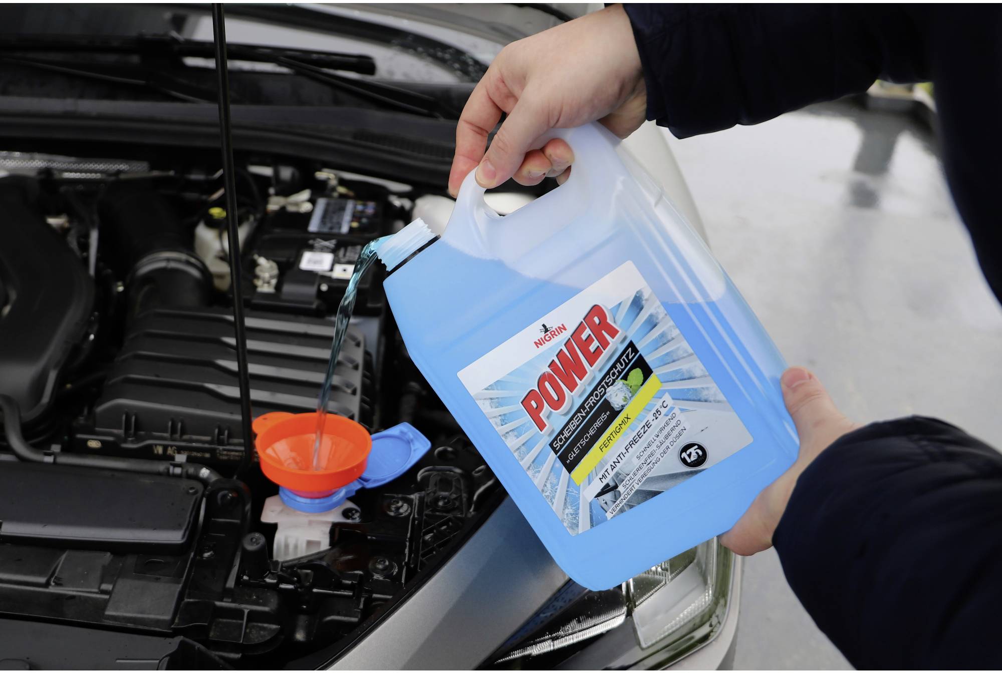 A person is pouring blue coolant from a plastic container into the engine bay of a car.