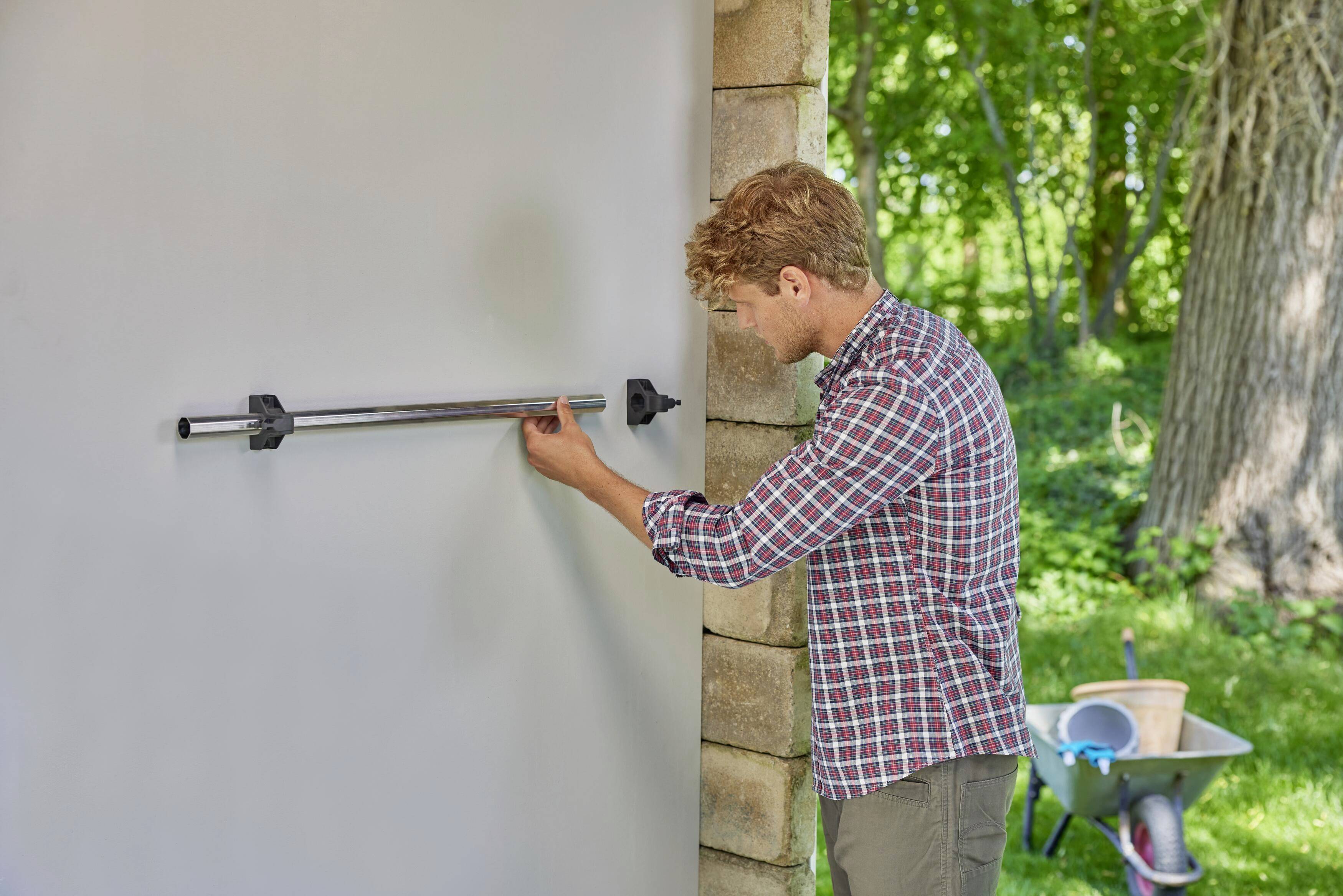 A man is installing a pole on a rectangular wall outdoors. A wheelbarrow is standing next to him. Trees can be seen in the background.