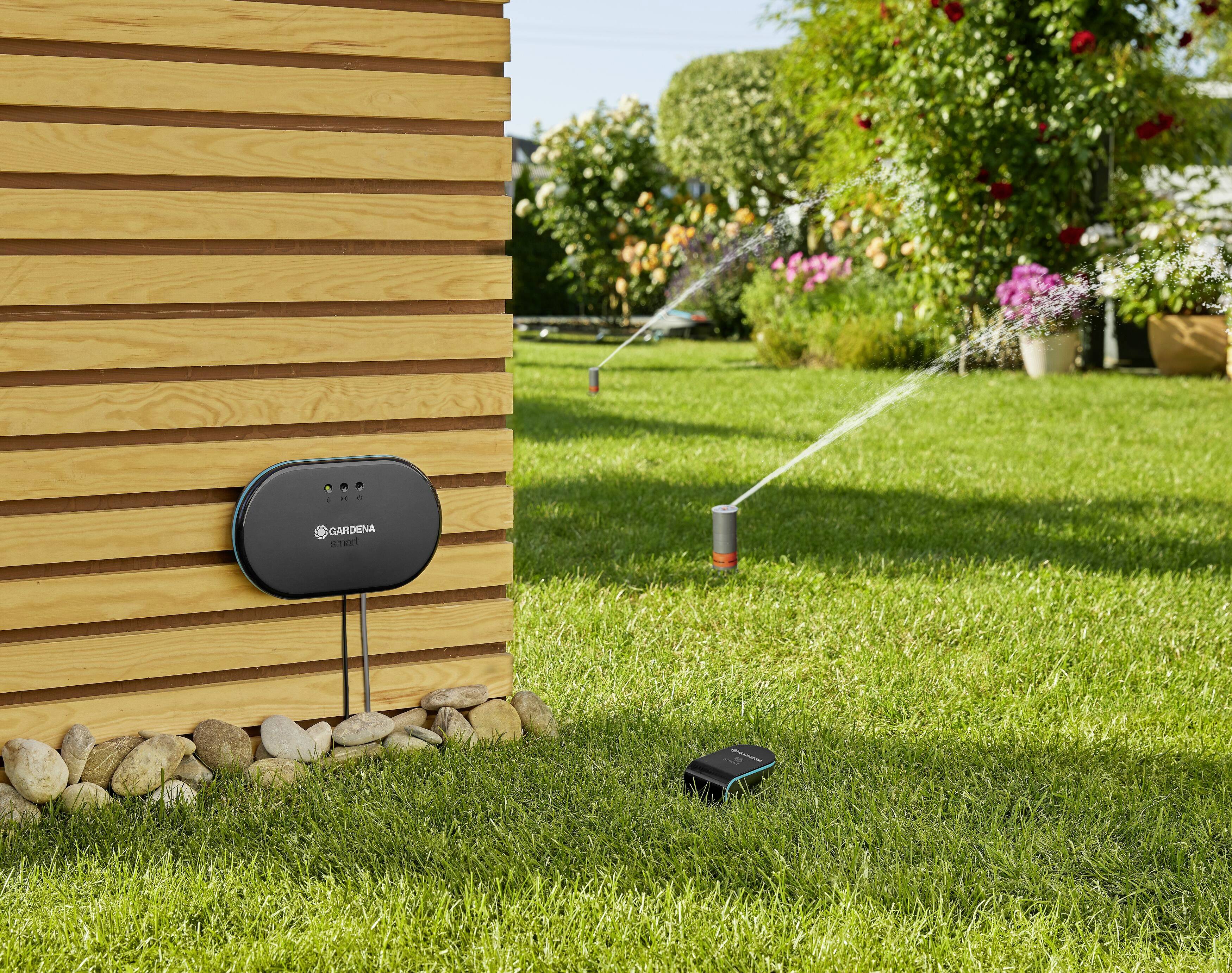 A garden irrigation system. Sprinklers on a green lawn, connected to a control unit on the house wall. Flowers in the background.