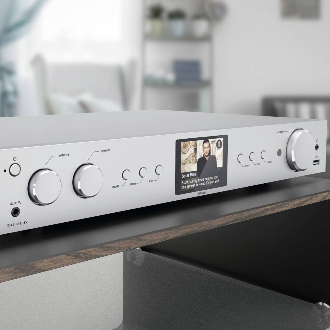 Silver amplifier with multiple controls and a small display showing a music track. A living room is visible in the background.