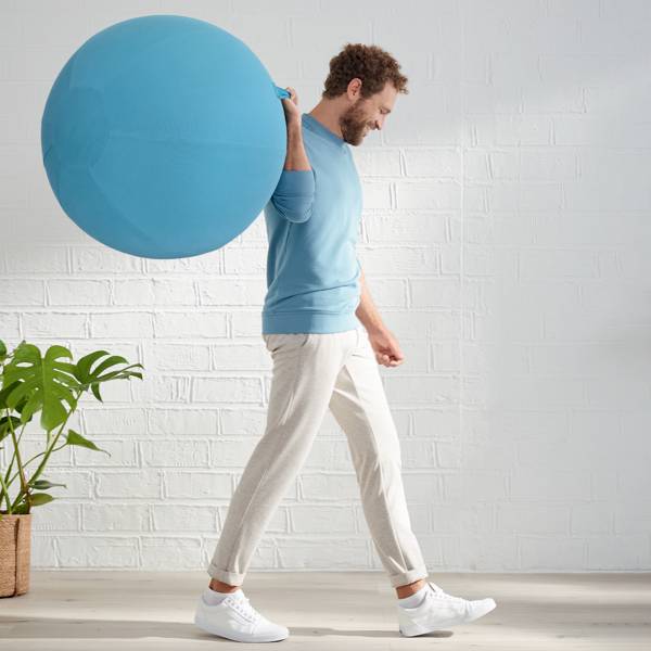 A man is carrying a large blue exercise ball and walking past a houseplant in front of a white brick wall.