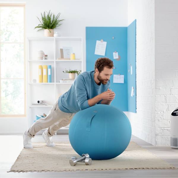 A man is doing planks on a large blue exercise ball in a modern, light-filled room, with a shelf in the background.