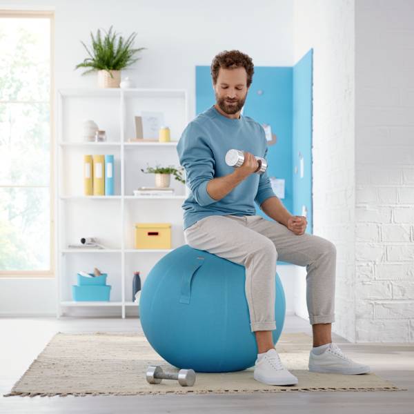 A man is sitting on an exercise ball in a living room and lifting a dumbbell. A bookshelf and a window are visible in the background.
