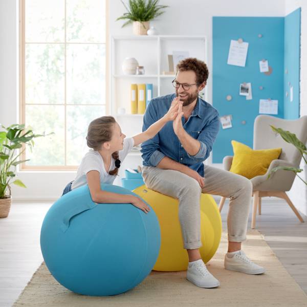 A man and a girl are sitting on colourful exercise balls in a bright room, clapping their hands. They are laughing and appear cheerful.