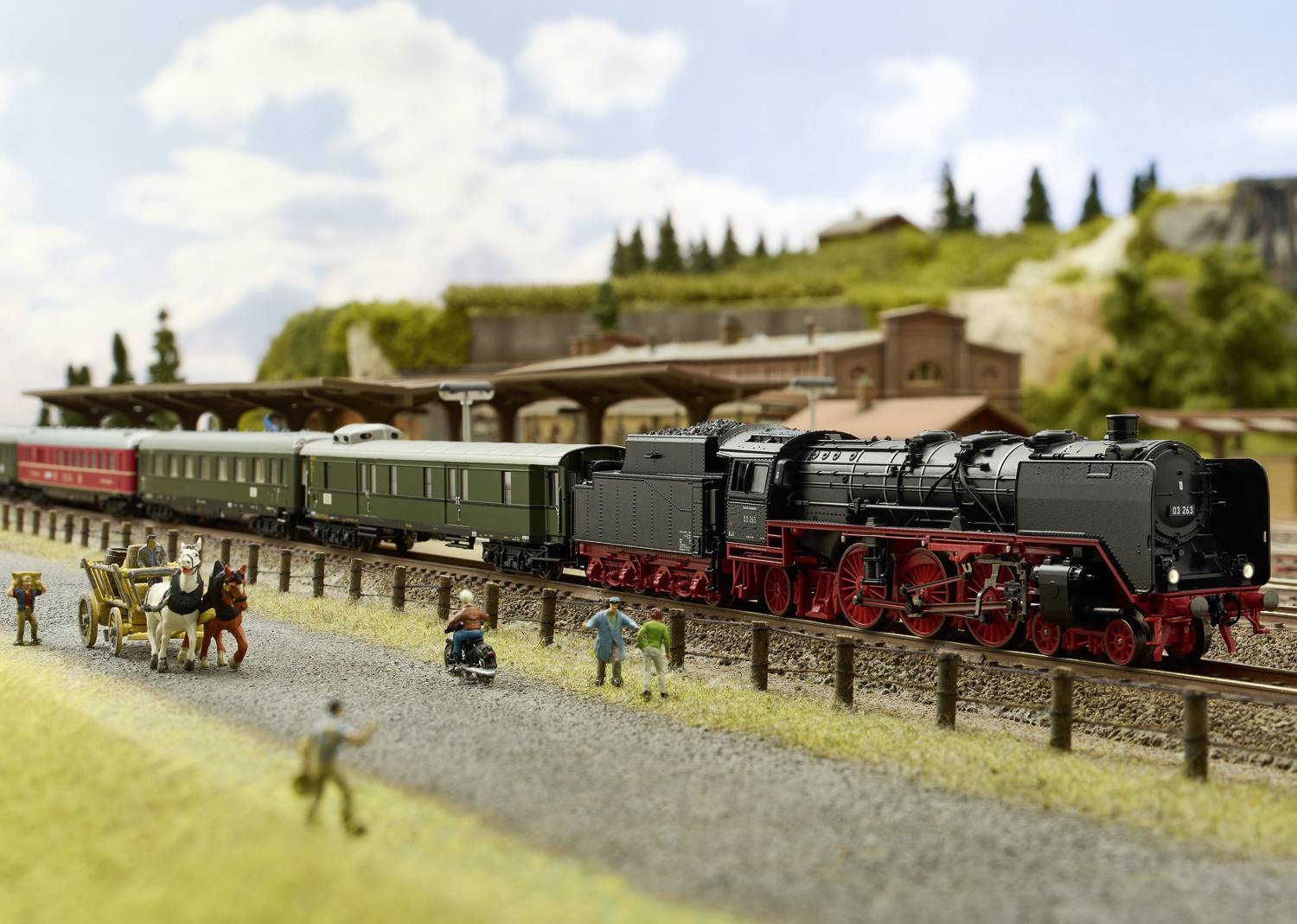 Model railway with a black steam locomotive and people on the platform. A horse-drawn carriage passes by, buildings in the background.