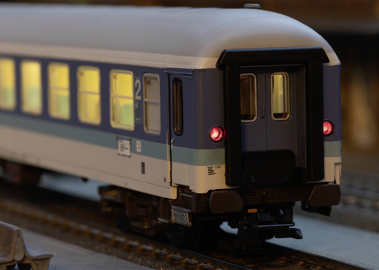 A model train with illuminated carriages on a track. The carriage is HO scale, blue and white, and bears the number '2' on its side.