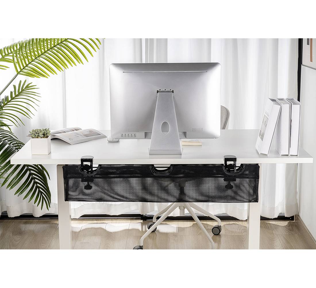 Desk with computer monitor, books and plants. A black mesh bag mounted underneath the table. Bright, modern environment.