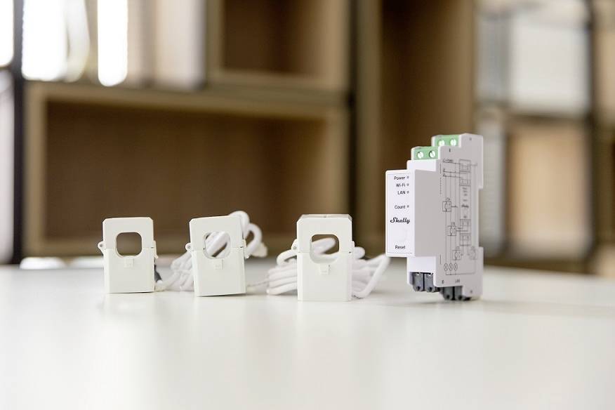 Three white, square sensors with cables are standing on a table next to a grey electrical device. Background blurred.