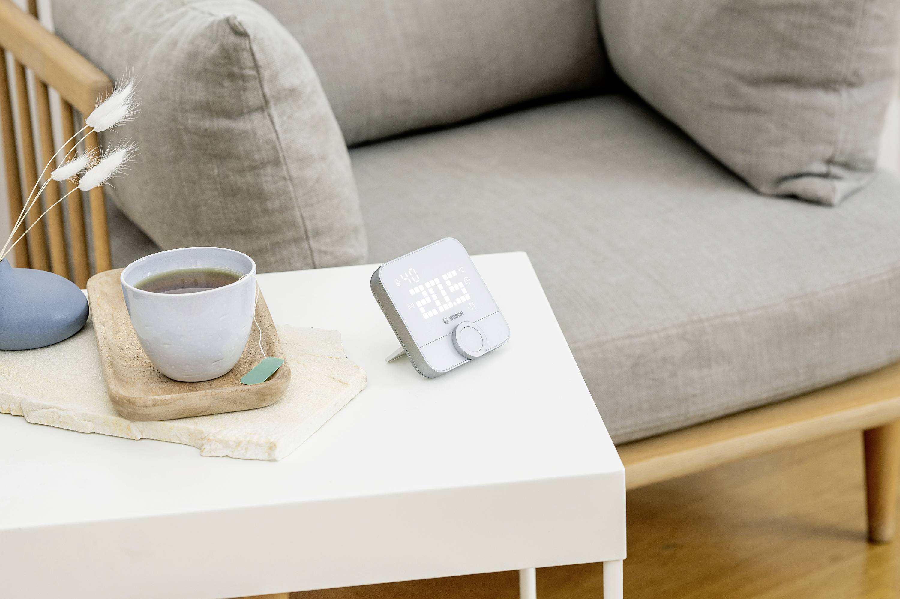A smart alarm clock with a white LED display shows '07:30'. It sits on a white table next to a cup of tea near a grey armchair.