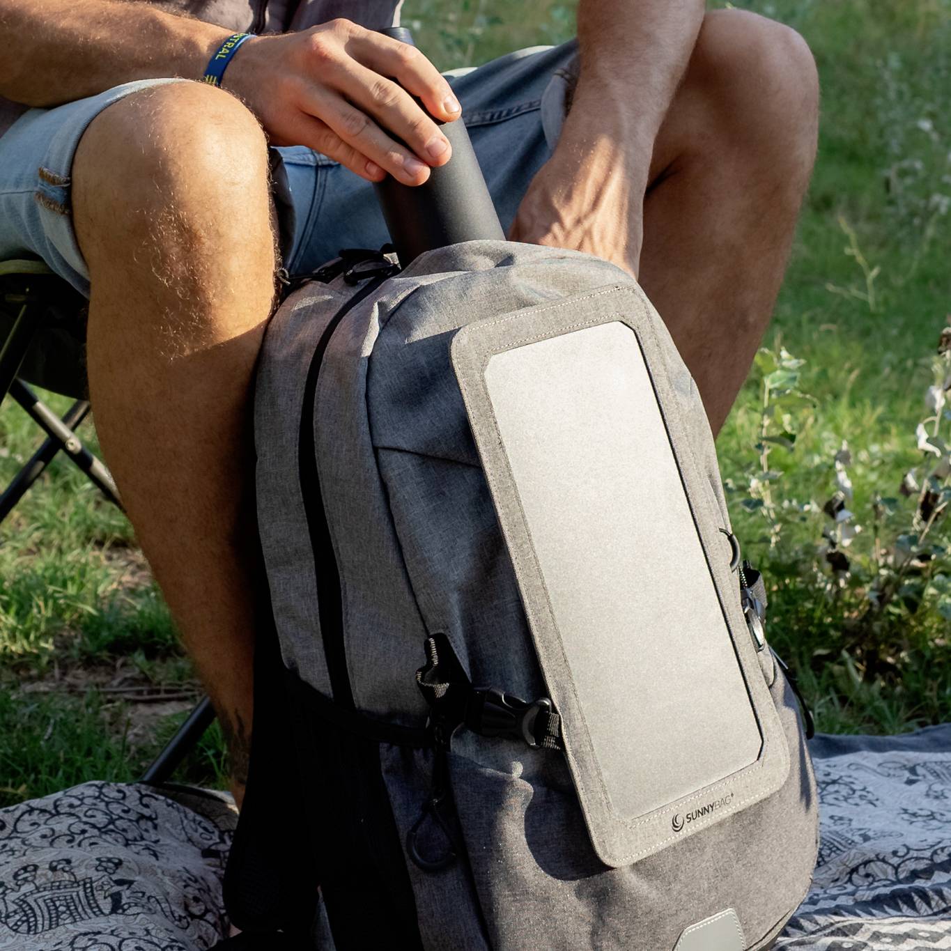 A person is sitting outdoors on a camping chair, holding a thermos flask, with a grey rucksack featuring a solar panel at the front.