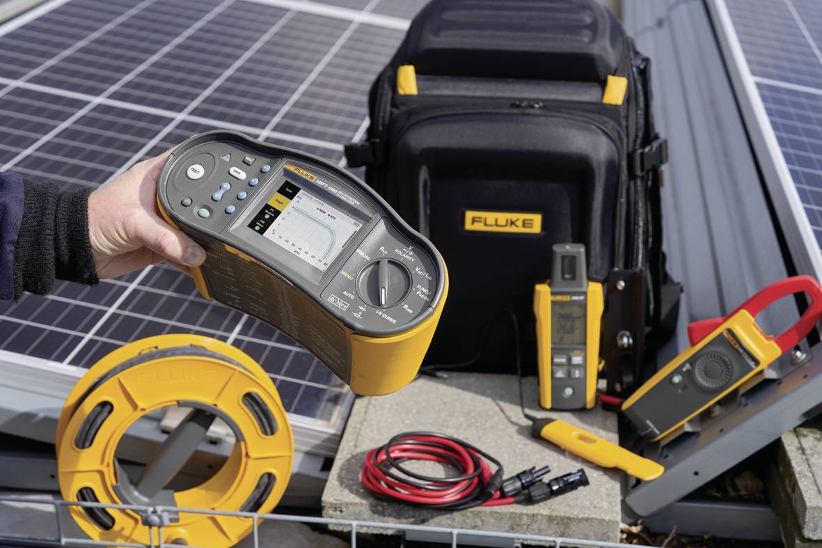 A person is holding a multimeter in front of a solar installation. In the background, additional measuring devices and a black tool bag are visible.