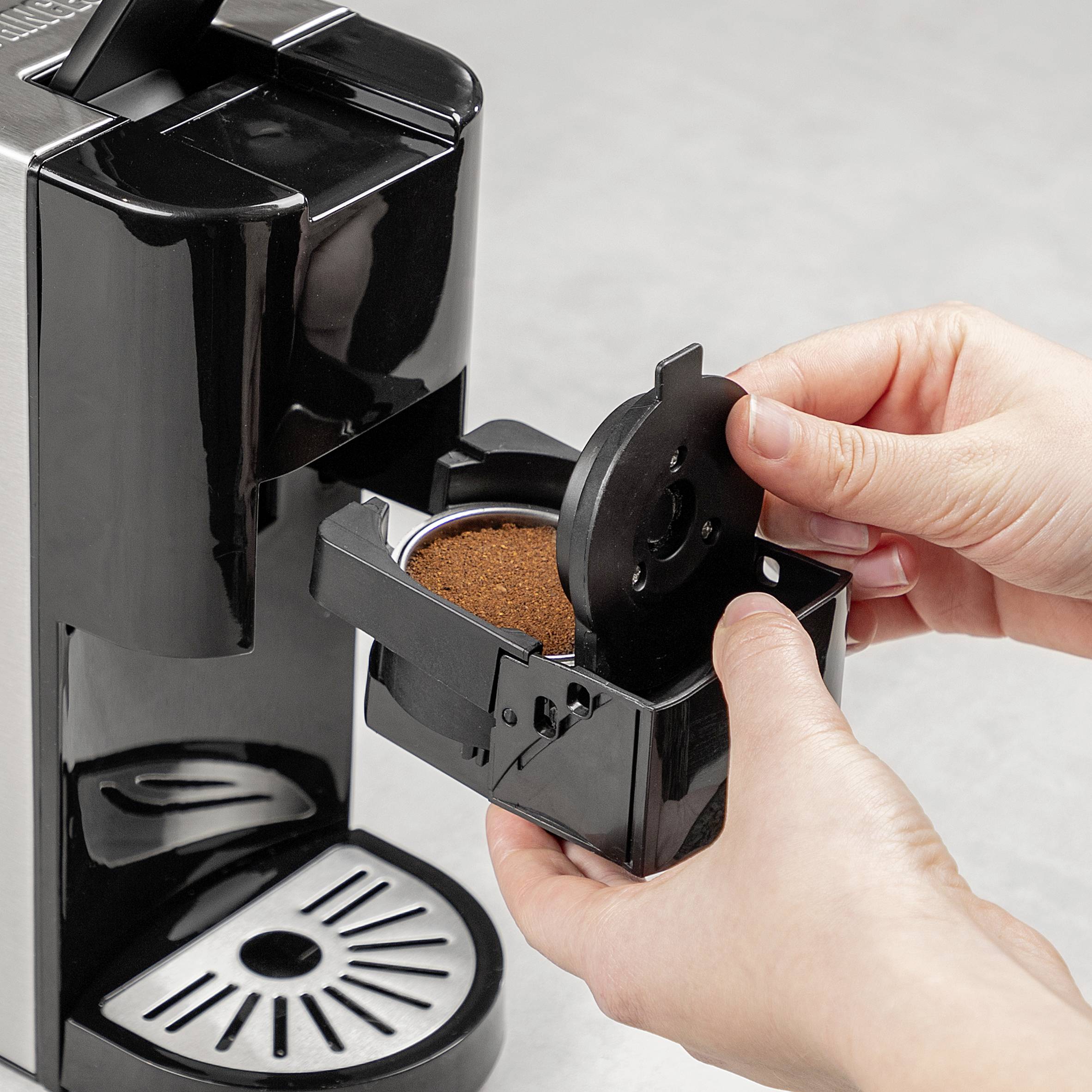 A person is filling coffee grounds into a capsule machine. The black machine is sitting on a grey surface.