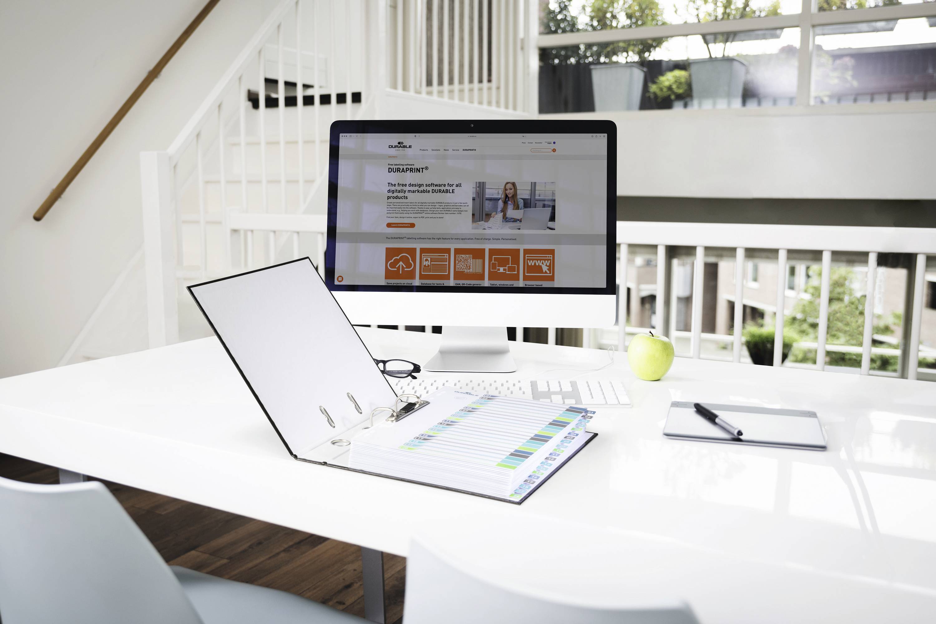 Modern workspace with a computer, ring binder, notepad and green apple on a white table. Window in the background shows a garden view.