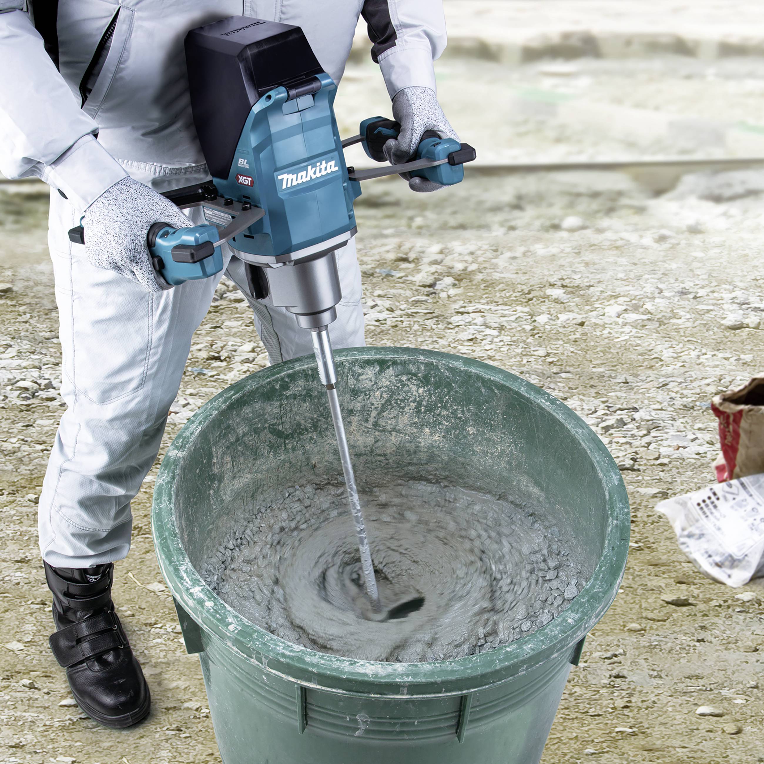 A person is mixing concrete in a green bucket using a Makita electric mixer. The person is wearing protective clothing.