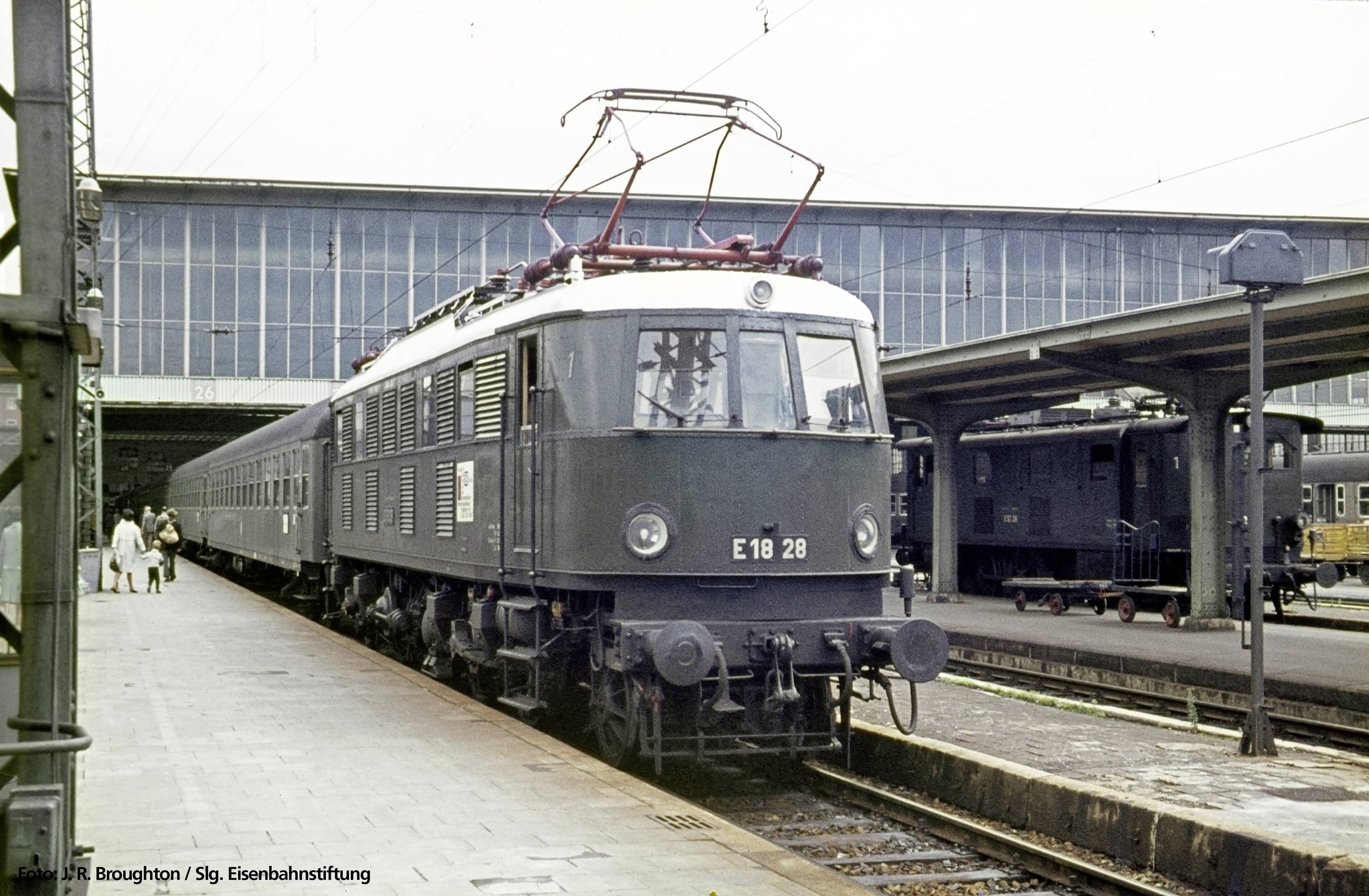 Train on a railway platform. A historic electric locomotive is pulling several passenger carriages. Buildings in the background with a glass façade.
