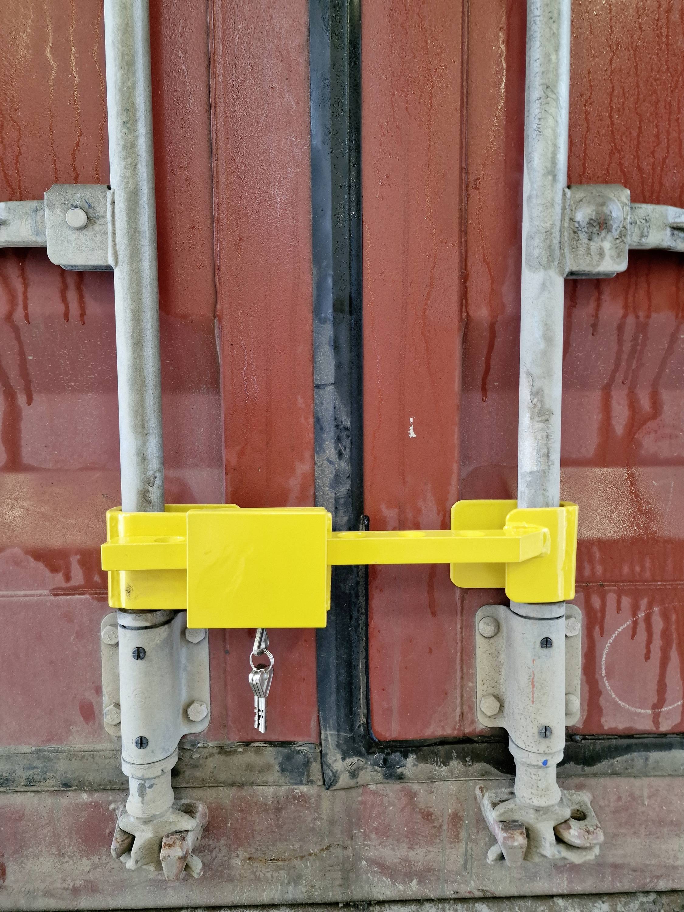 A yellow security padlock secures the closed doors of a red freight container. A key is hanging on the lock.