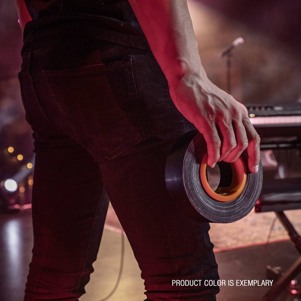 A person on a stage is holding a roll of tape. In the blurred background, a keyboard and microphone are visible.